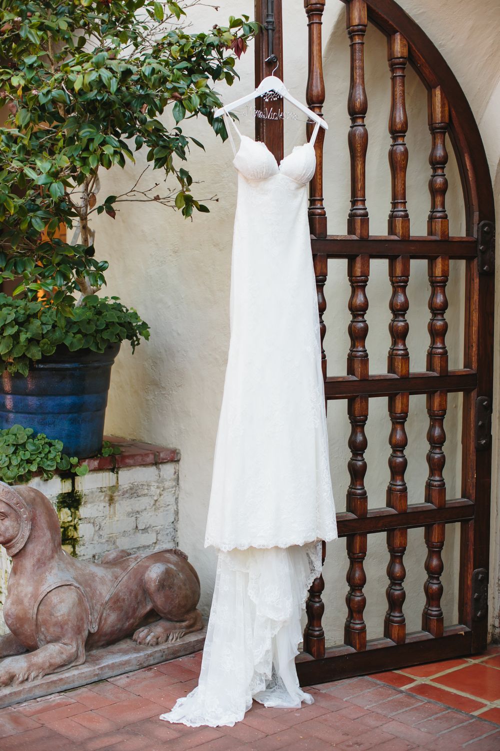 White lace wedding dress hanging against a rustic wooden archway with decorative balusters and green foliage.