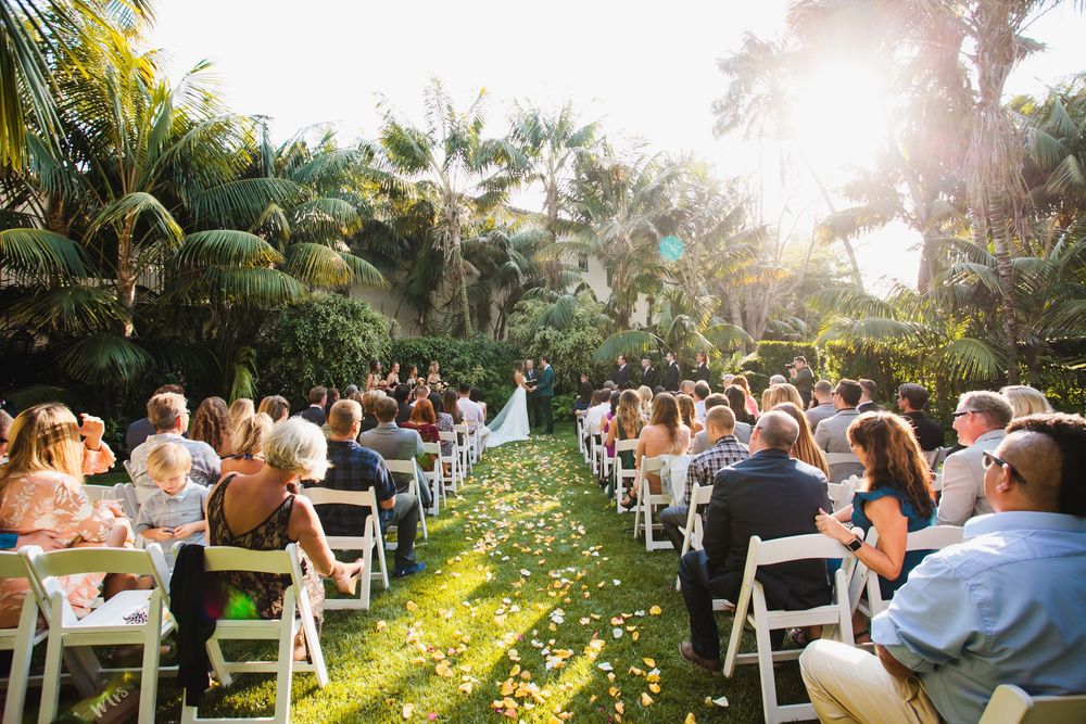 Guests seated on white chairs for an outdoor garden wedding ceremony with tropical palm trees and golden sunlight.