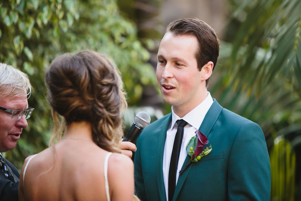 Wedding party in dark green suits and formal attire gathered outdoors among tropical plants.