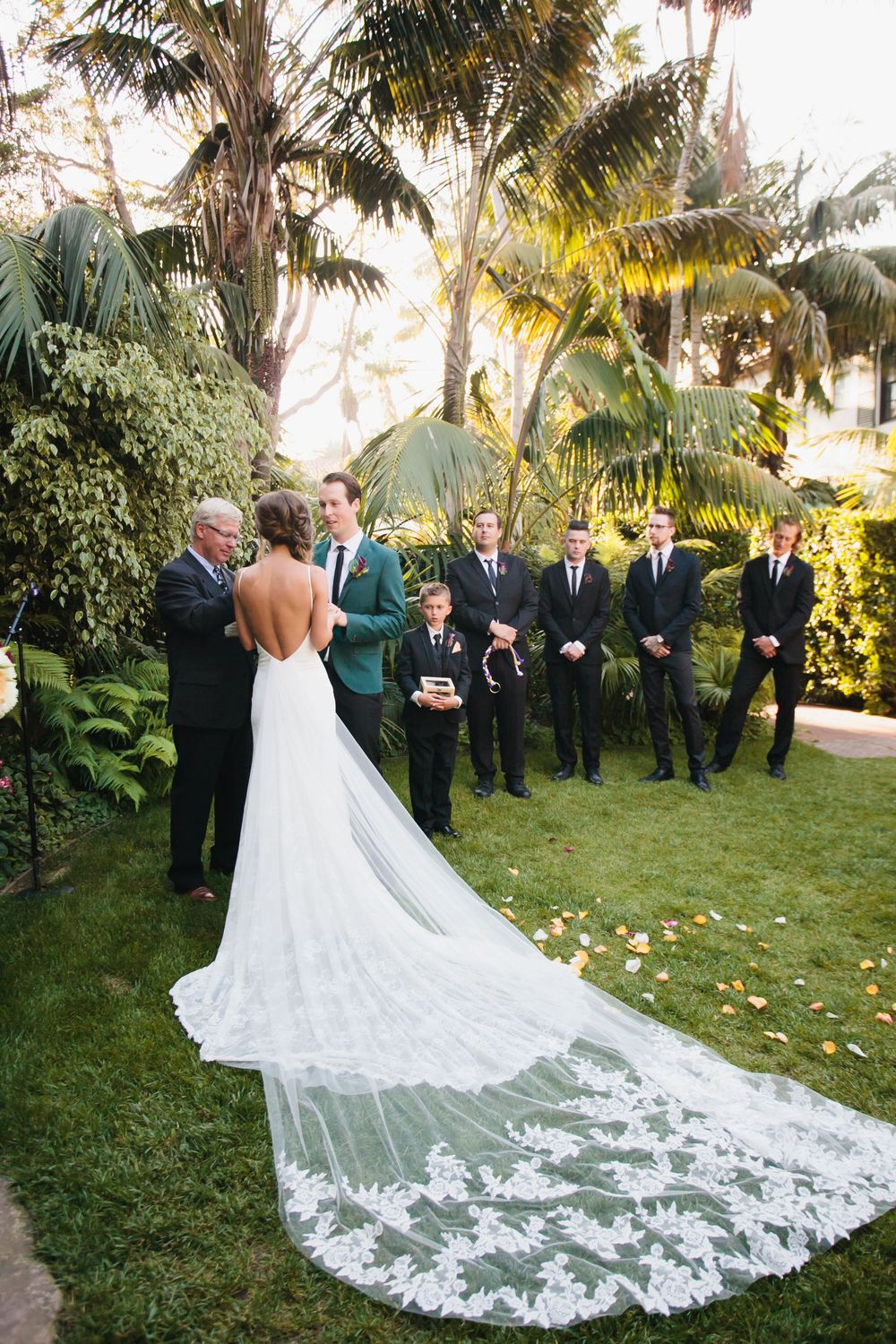 A bride in a backless gown with long lace train poses with groomsmen in an outdoor garden setting.