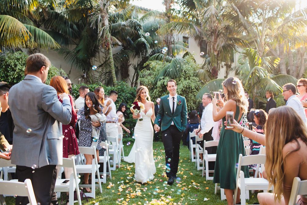Outdoor wedding celebration with guests gathered among palm trees at sunset.