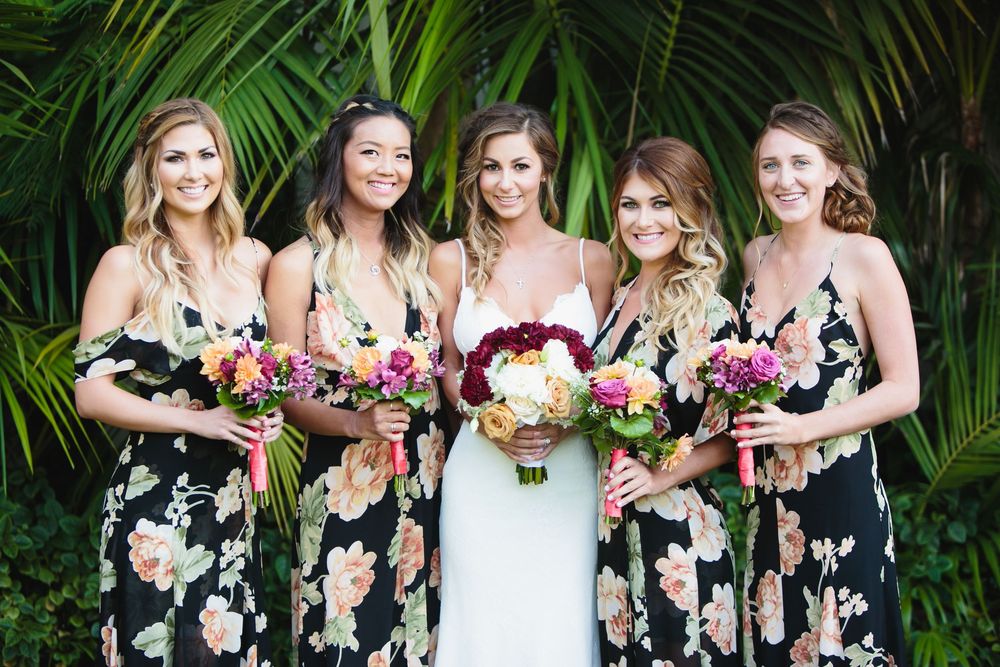 Bridal party in floral black dresses poses with bouquets against tropical foliage.