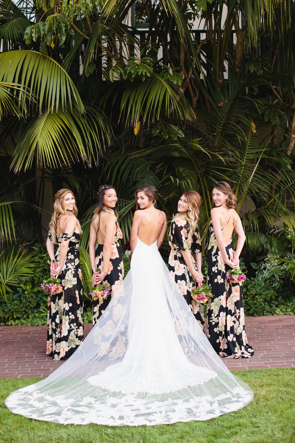 Bride in lace gown with bridesmaids in floral dresses showcasing the train against palm trees.