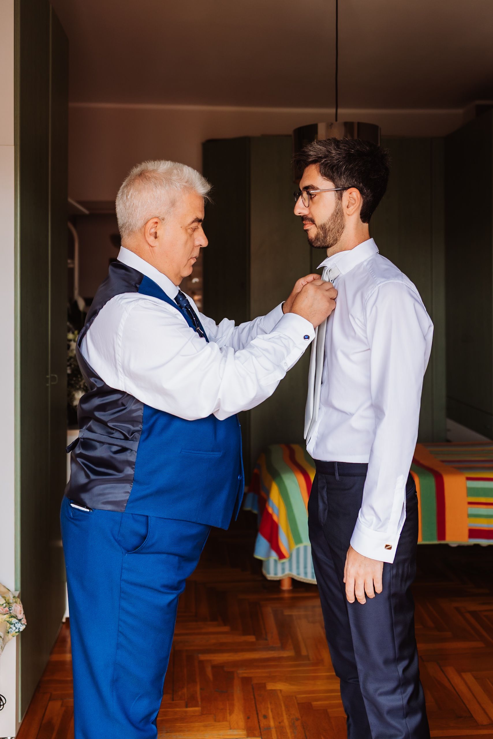 Two people in formal attire help each other with adjusting a tie in an indoor setting.
