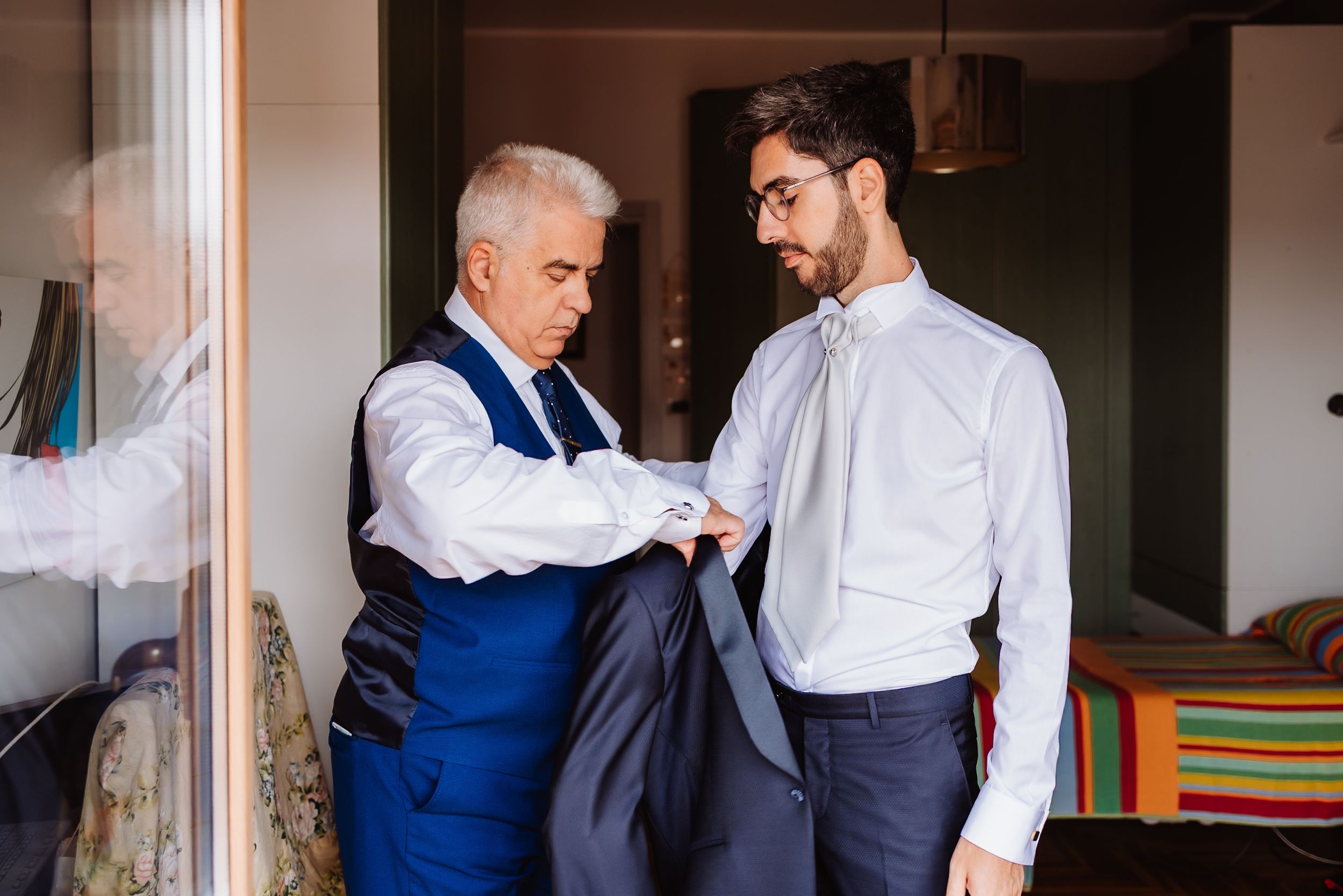 Two men in formal attire help each other get dressed in an indoor setting.
