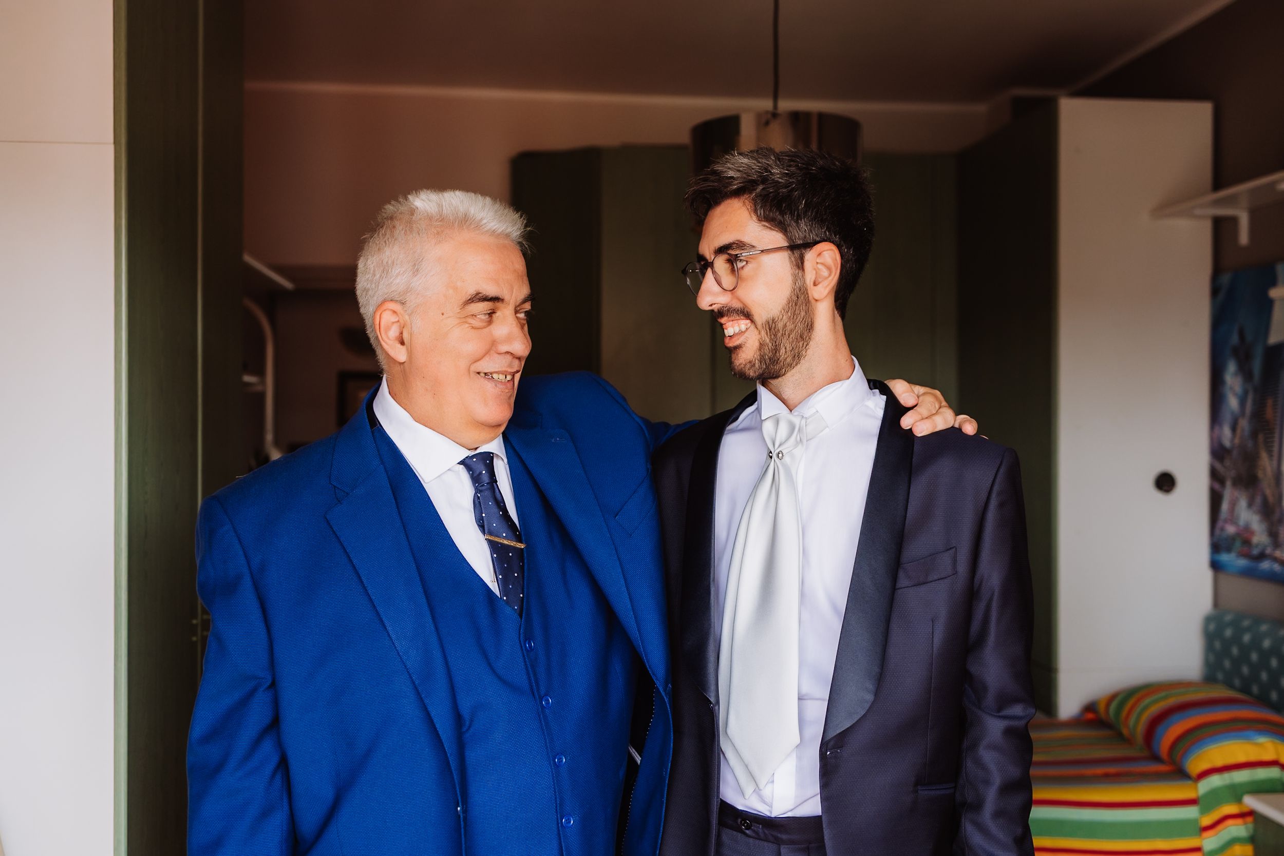 Two men in blue and navy suits share a moment while getting ready indoors.