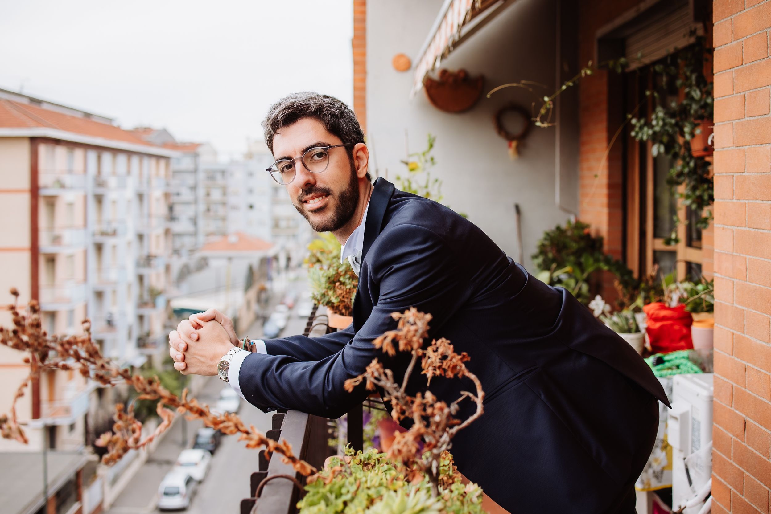 Two men in formal wear stand on a balcony with brick walls and potted plants.