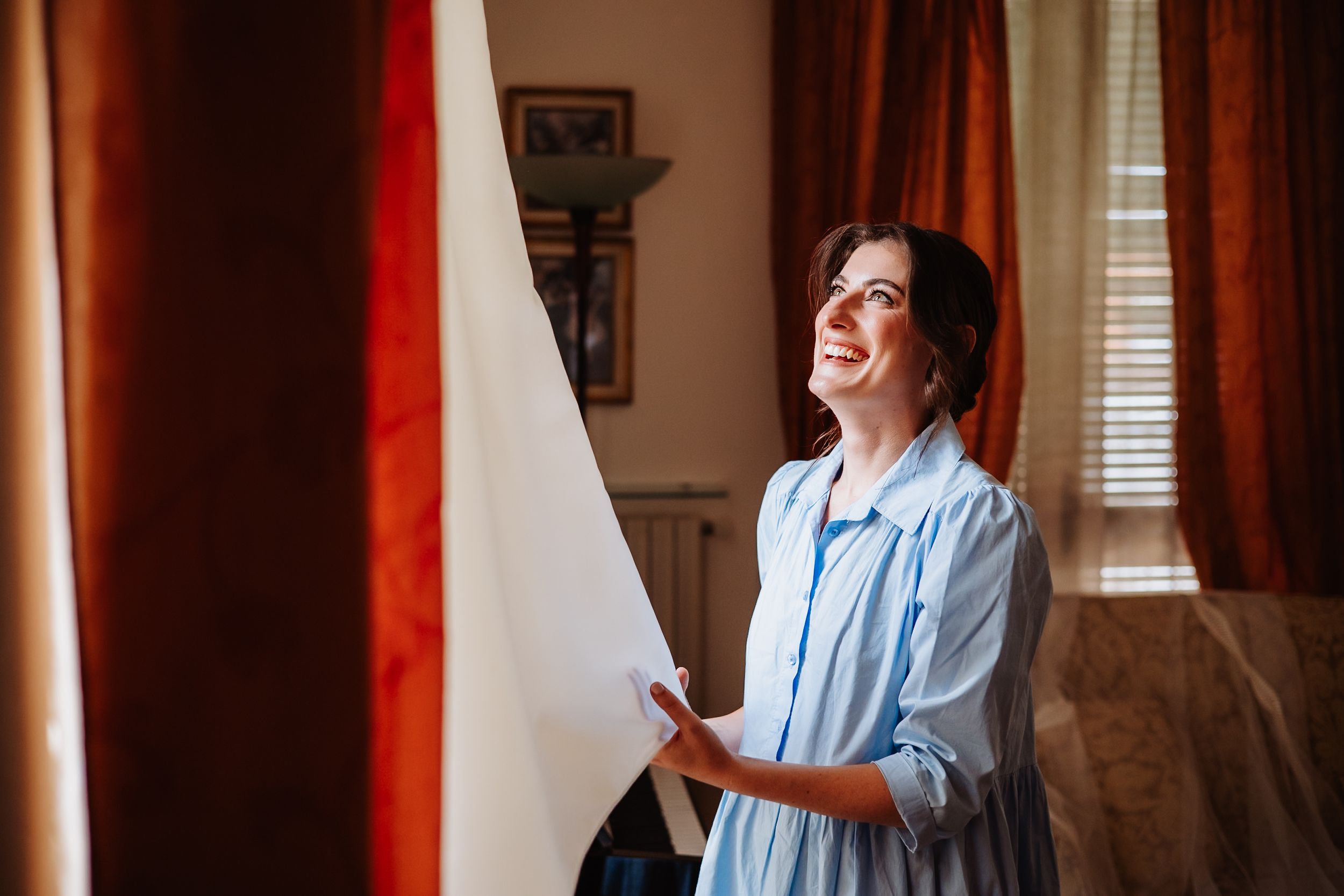 Woman in light blue dress standing by window with red curtains looking up in contemplative pose.