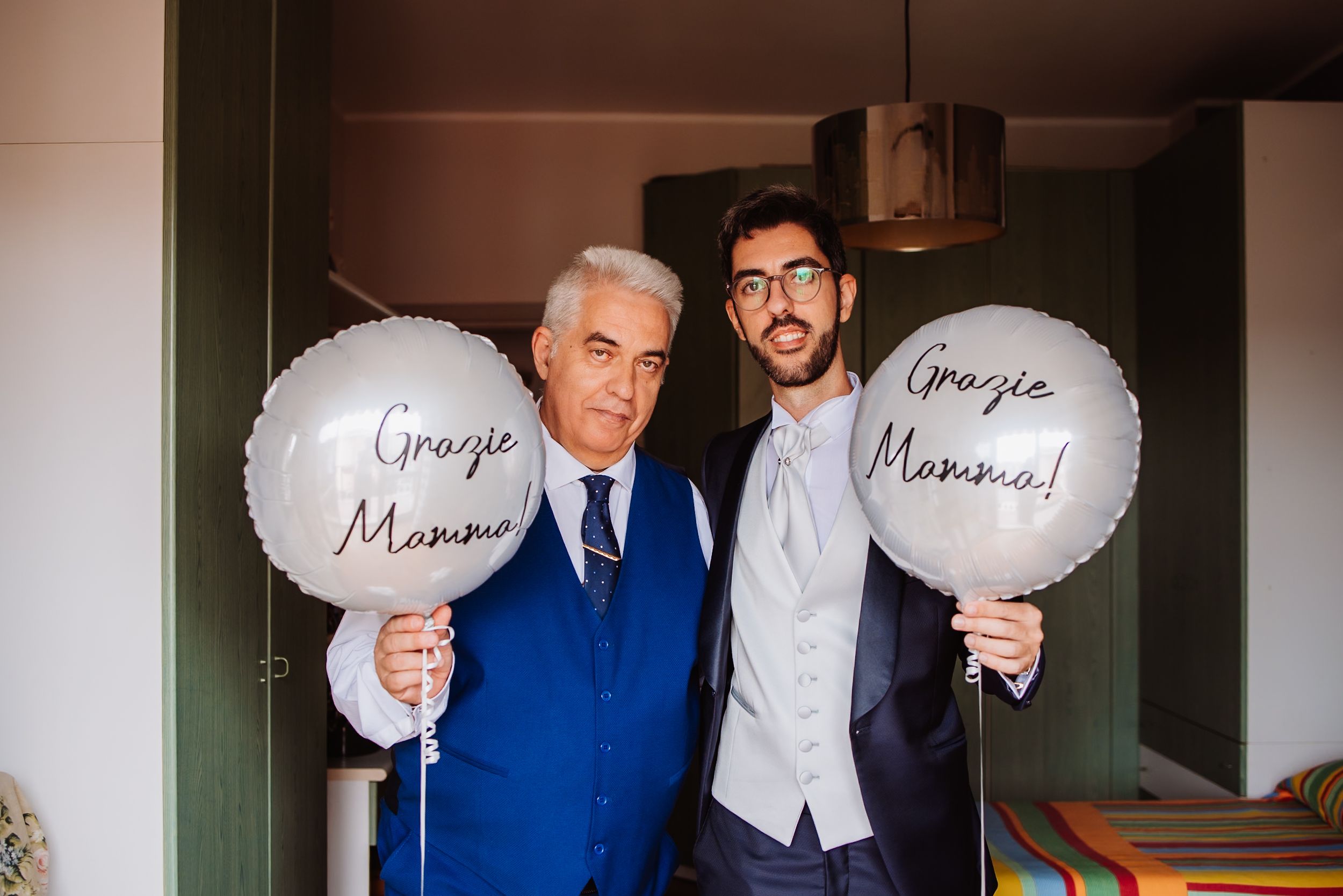 Wedding party members hold white balloons with text while posing together in formal attire.