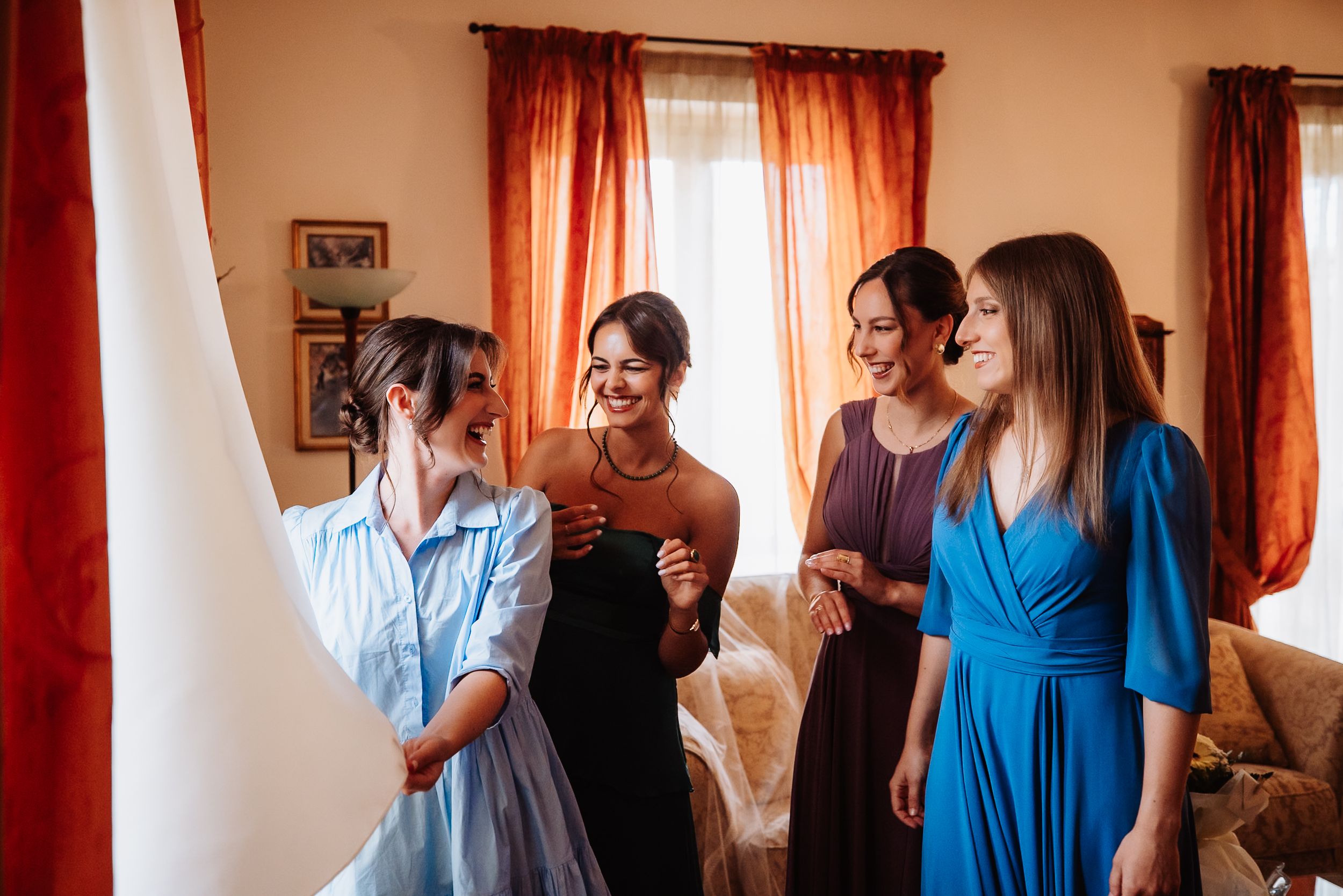 Group of people in blue and dark dresses gathered in room with red curtained windows.