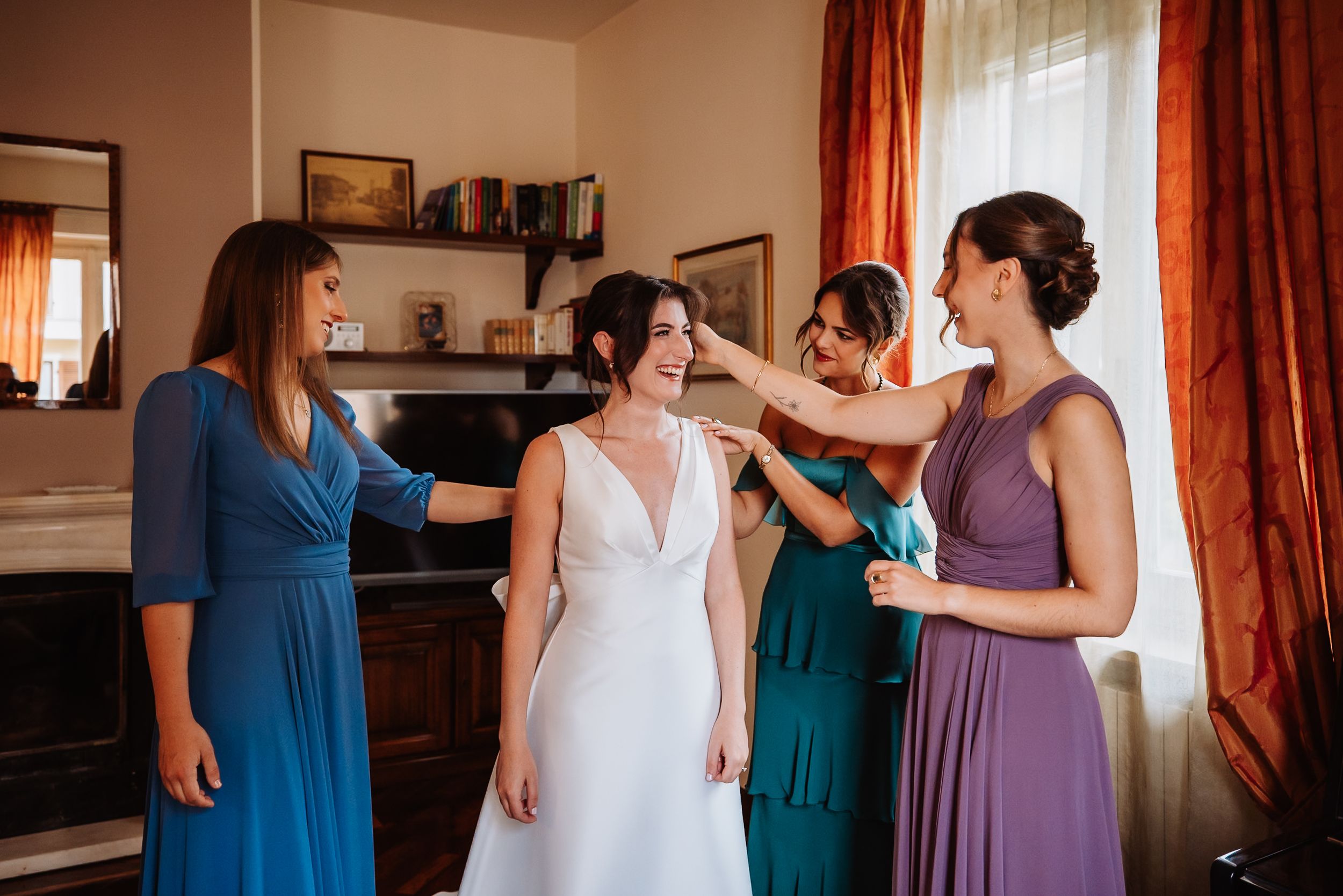 Bridesmaids in coordinated blue and purple dresses help the bride get ready in a sunlit room.