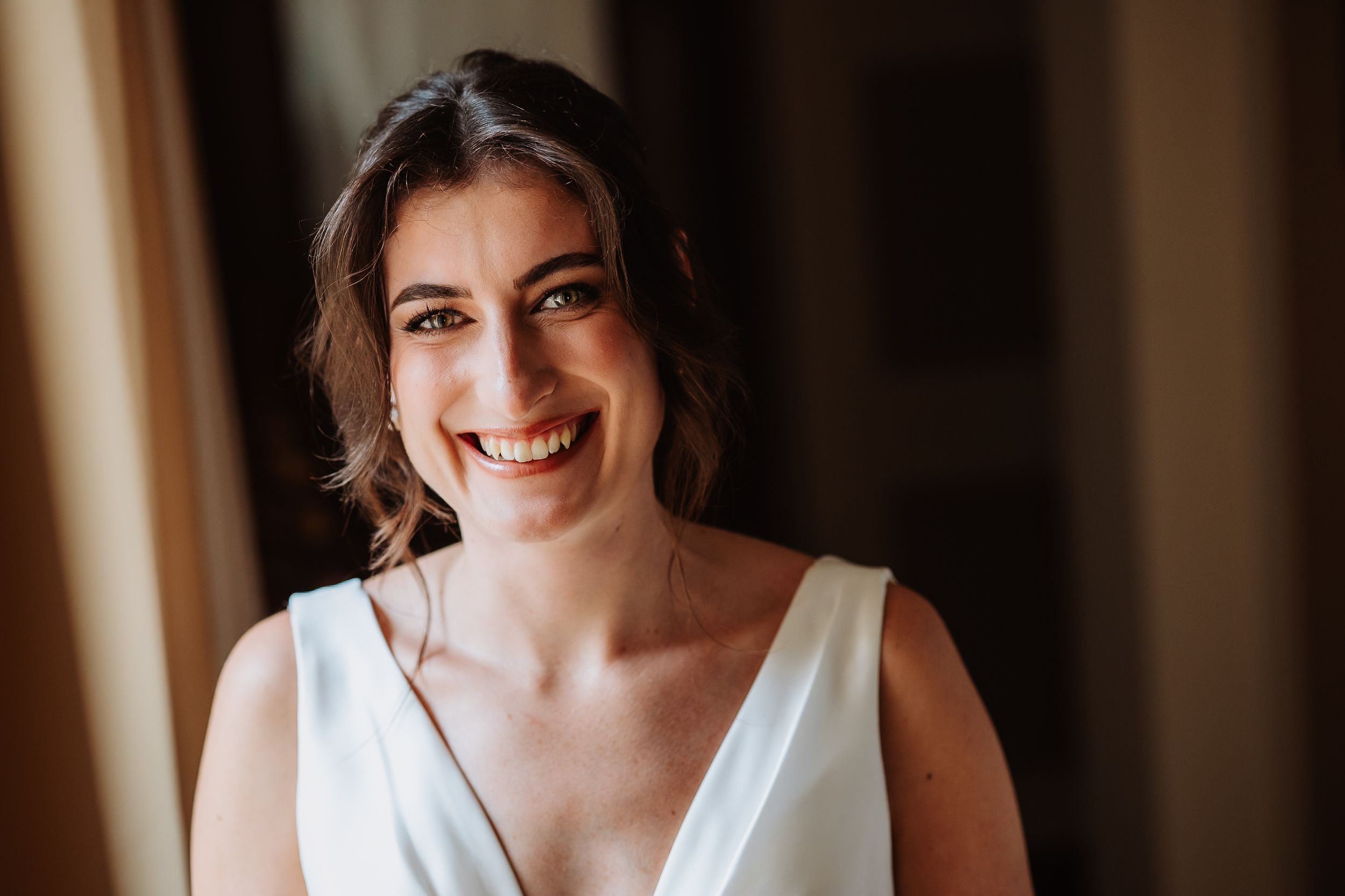 A joyful bride wearing a white V-neck wedding dress smiles warmly in natural window light.
