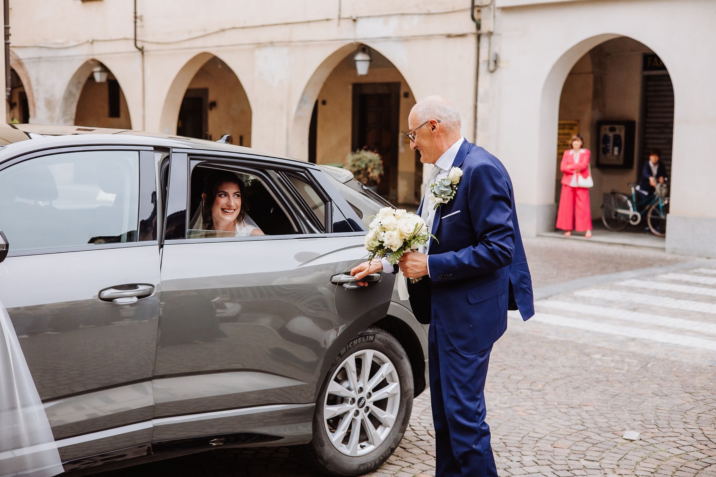 A person in a navy blue suit stands by a gray car holding flowers while someone sits in the back seat.
