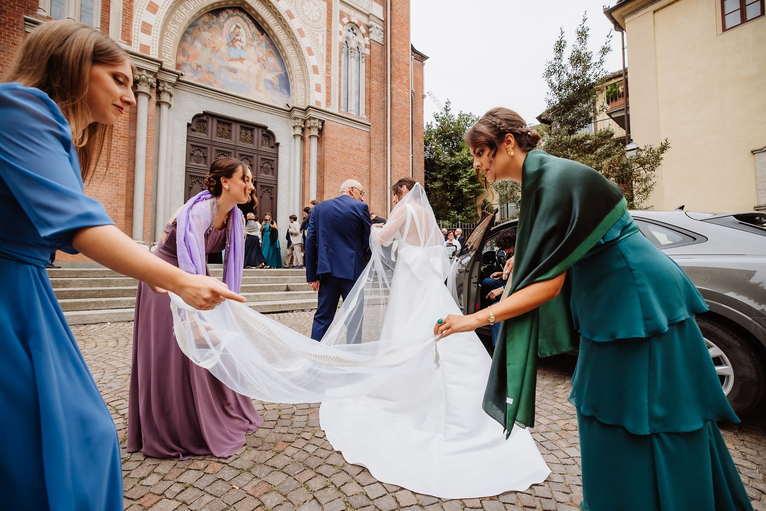 A group helps adjust a flowing white wedding veil on a cobblestone street near historic buildings.