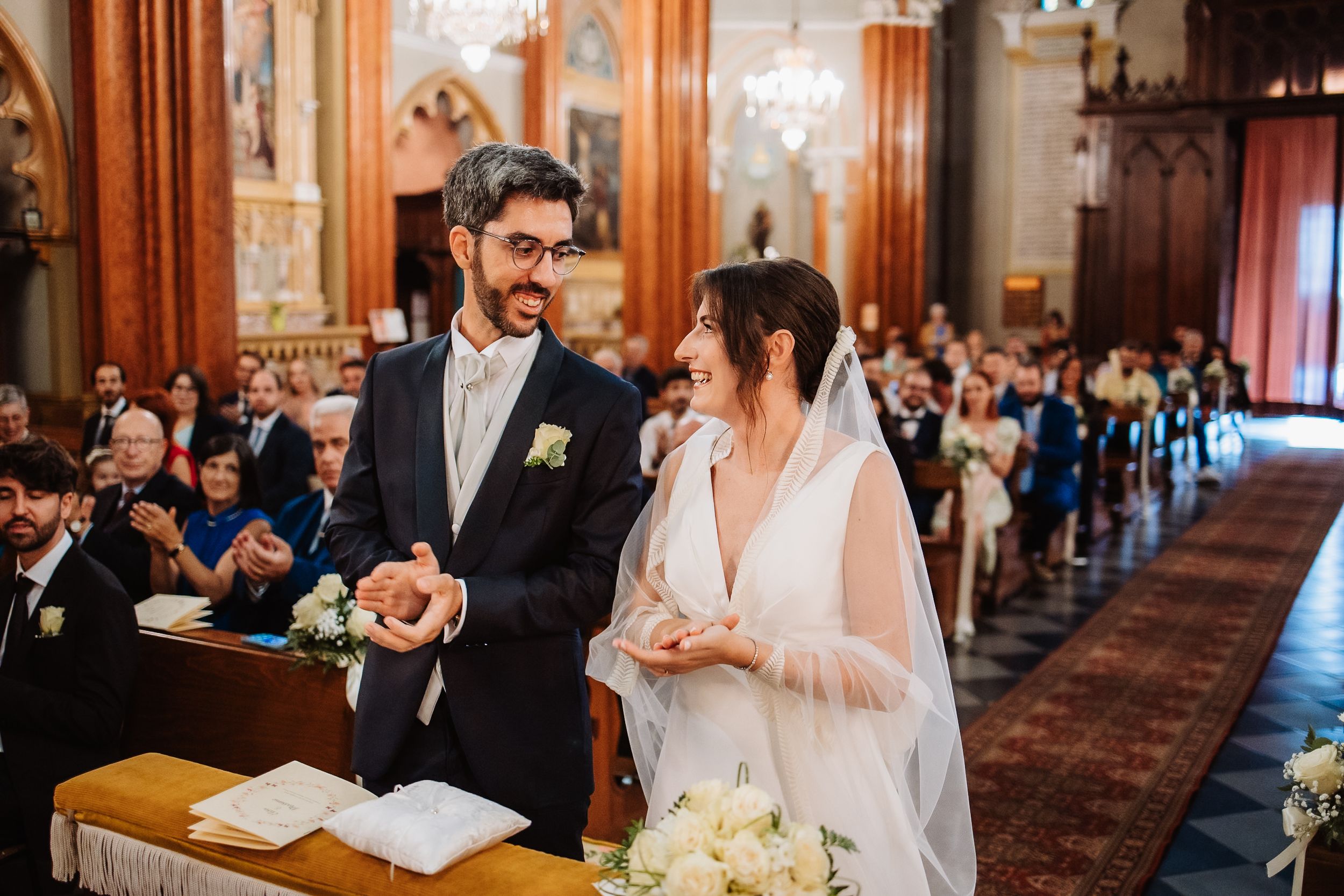 A couple exchanges rings during their wedding ceremony inside an ornate church with stained glass windows and chandeliers.