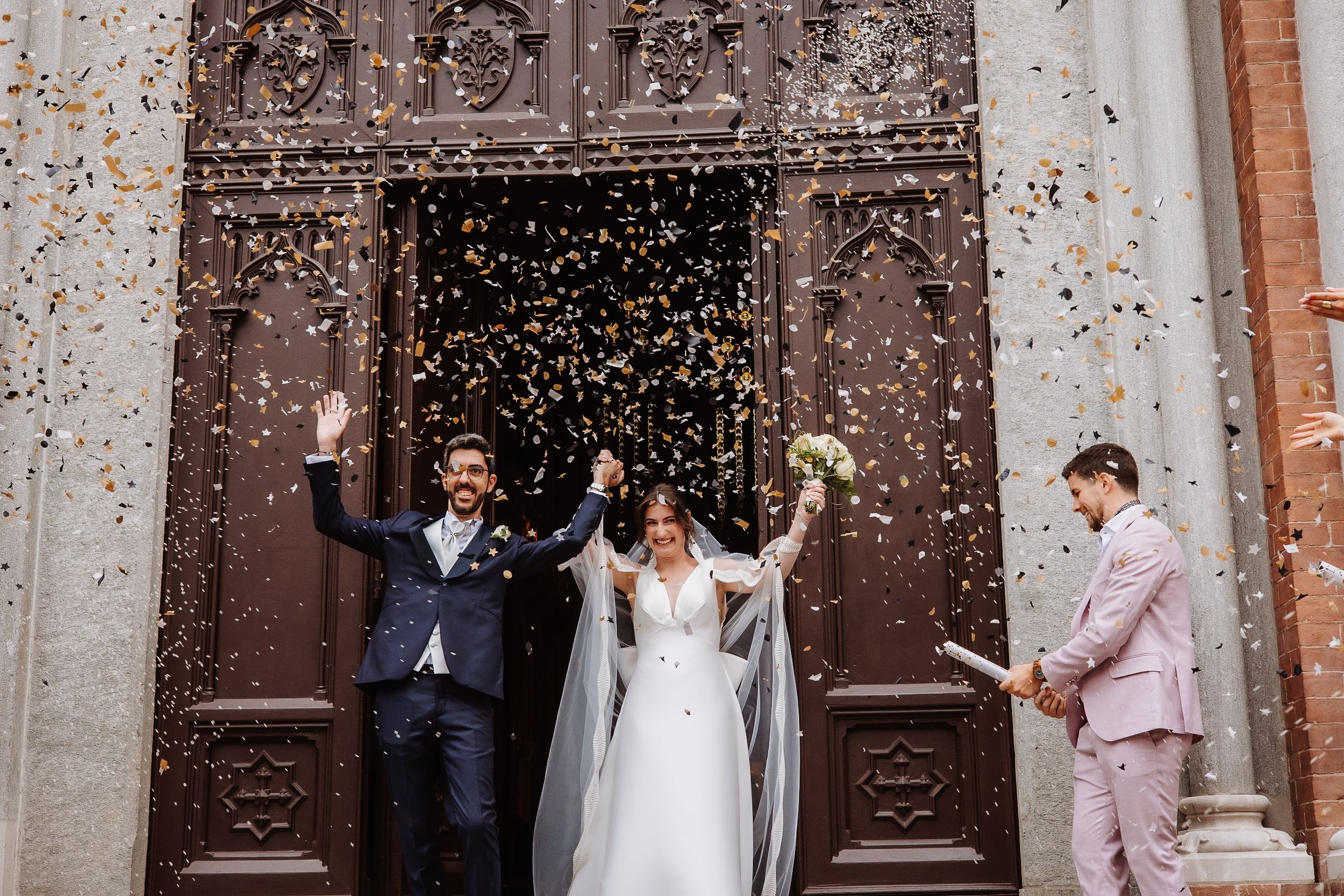 Wedding guests throw confetti at a newlywed couple outside an ornate stone church entrance with wooden doors.