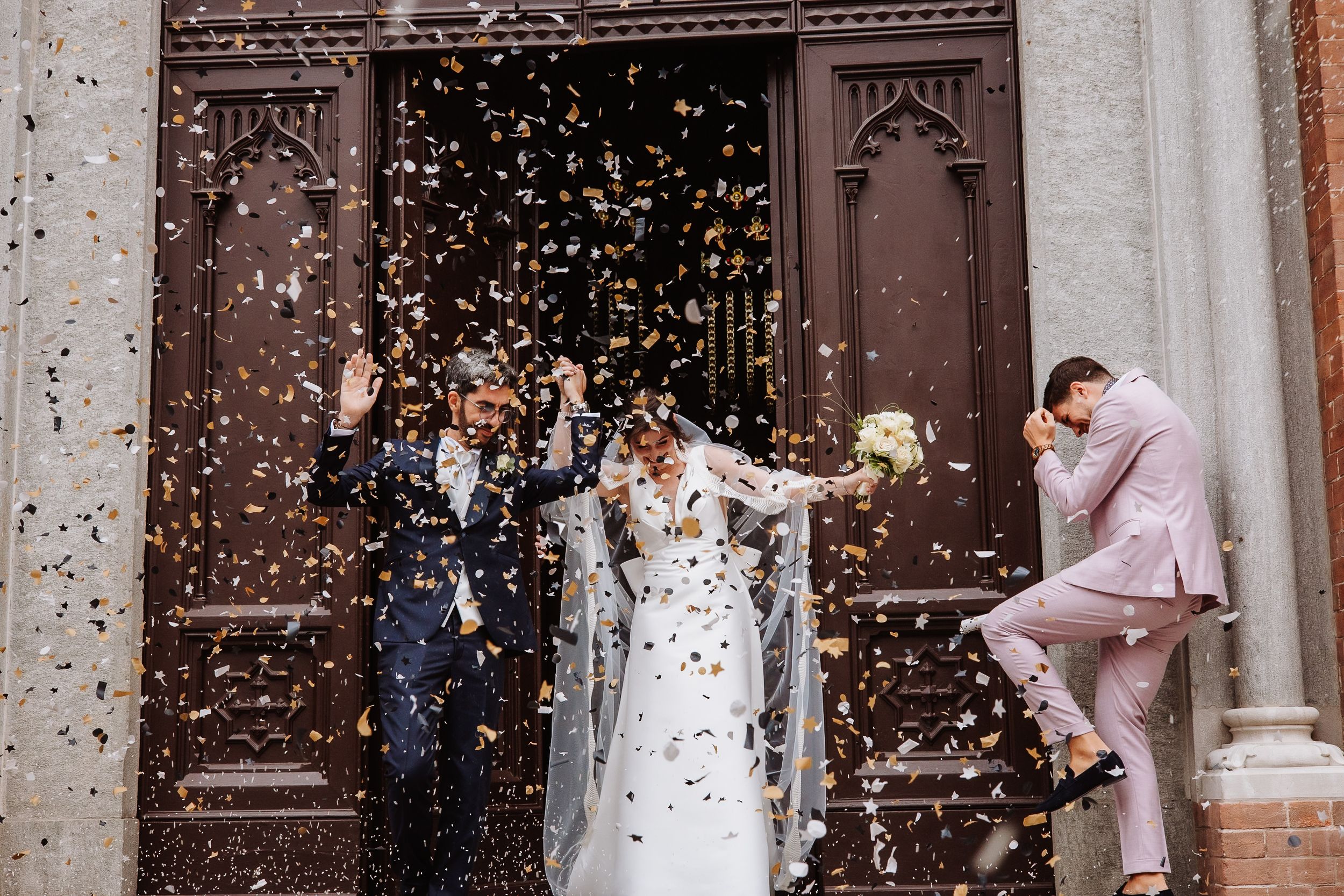 Wedding guests throw white confetti over a bride and groom as they exit a traditional church building.
