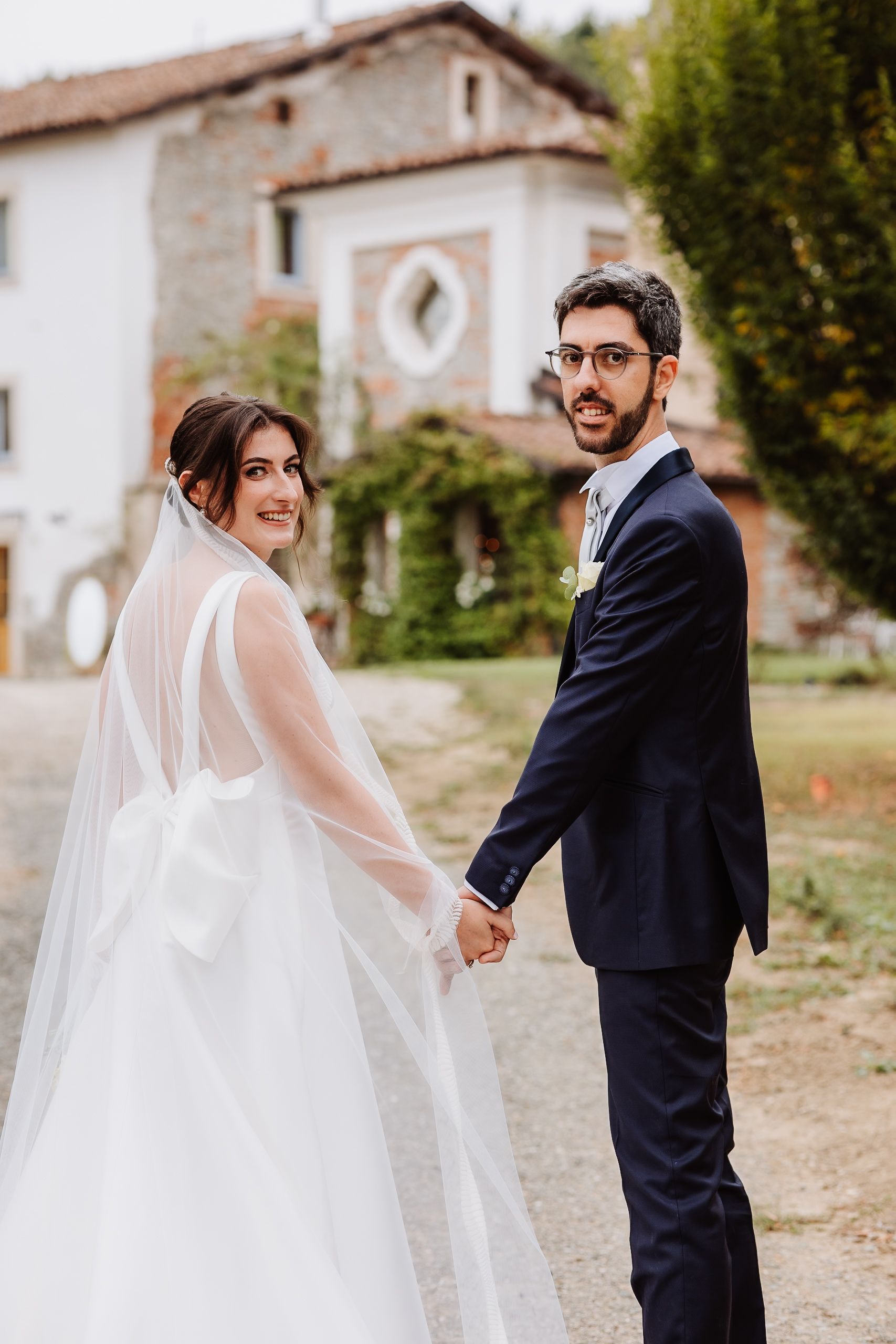 A couple in wedding attire share candid moments in front of a rustic white building on their special day.
