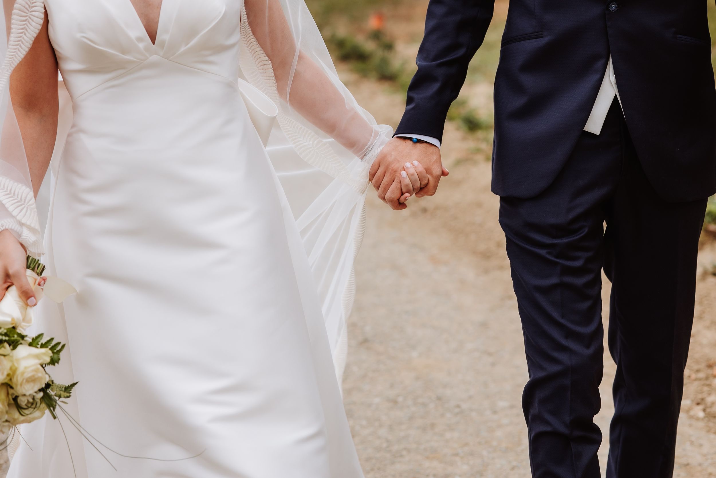 A couple in wedding attire walking hand in hand on a gravel path during an outdoor ceremony.
