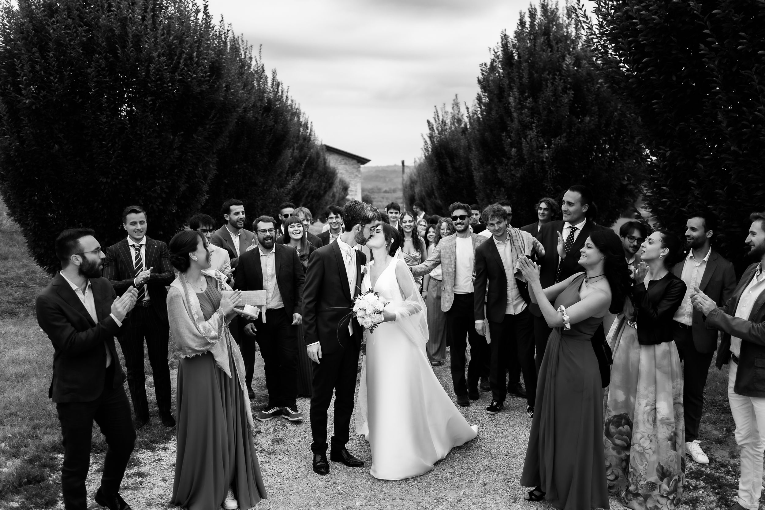 A black and white wedding photo showing a group of guests gathered on a tree-lined path during an outdoor celebration.