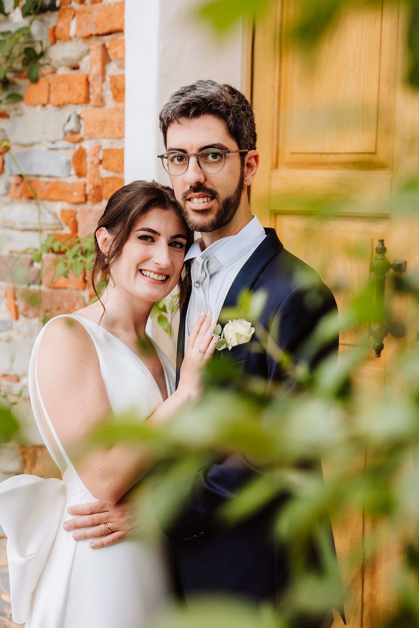 Newlywed couple shares romantic moments against rustic brick wall with green foliage in foreground.