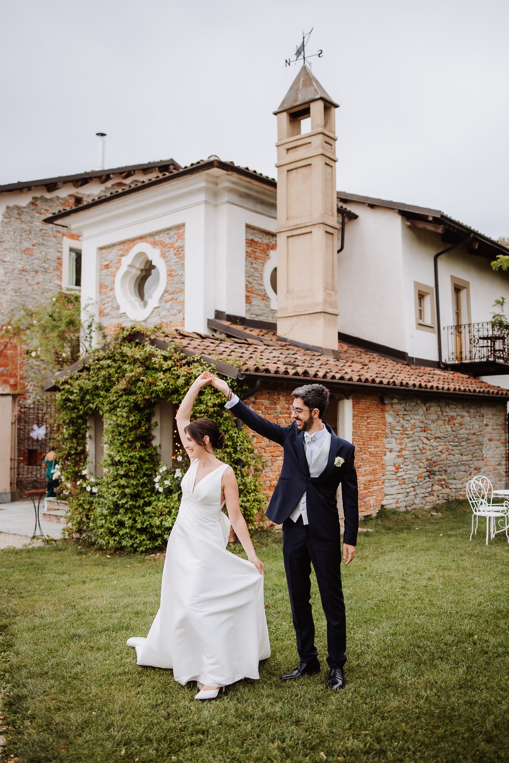 Bride and groom share playful moments dancing and twirling on a grassy lawn beside a rustic stone building.