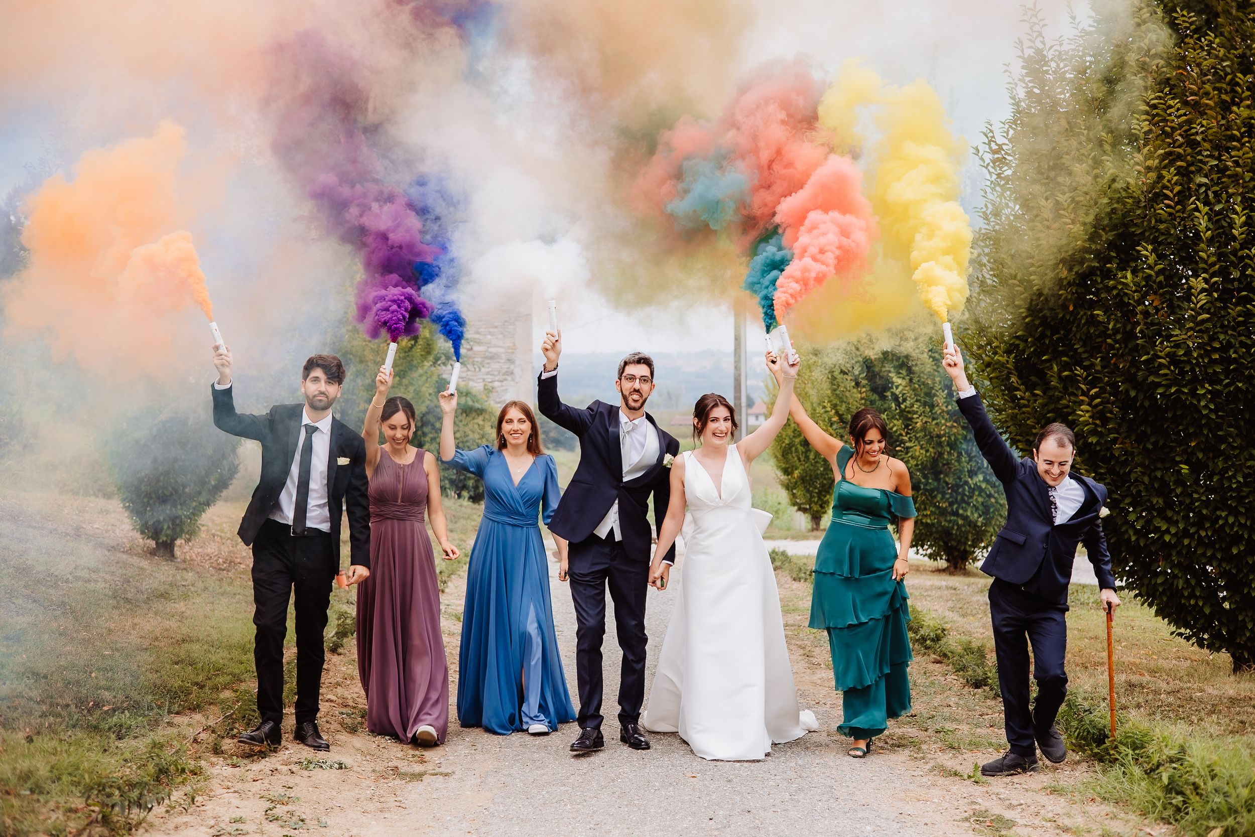Wedding party celebrates with colorful smoke bombs in an outdoor setting during golden hour.