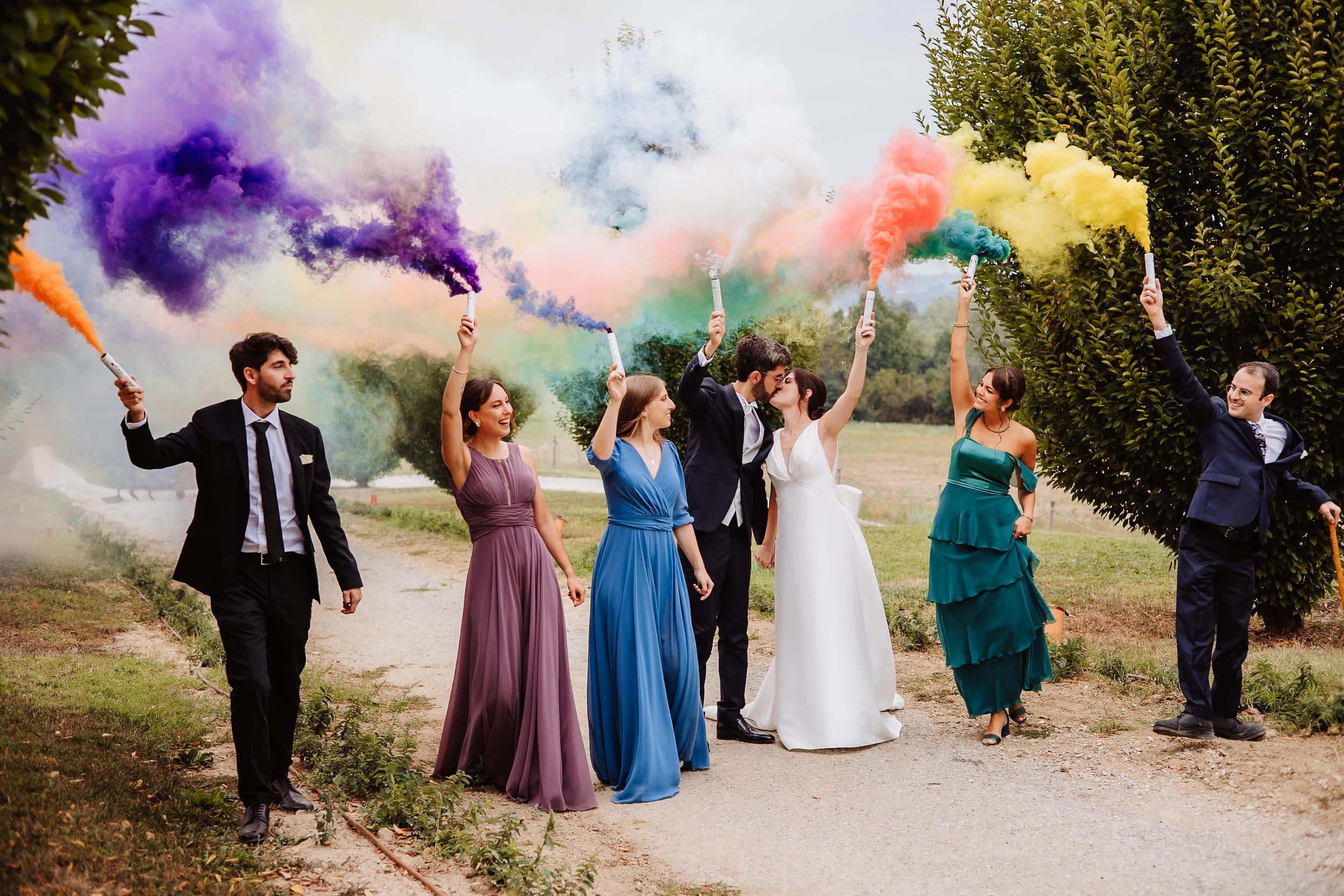 Wedding party celebrates on a path with colorful smoke bombs creating vibrant purple, blue, pink and yellow clouds.
