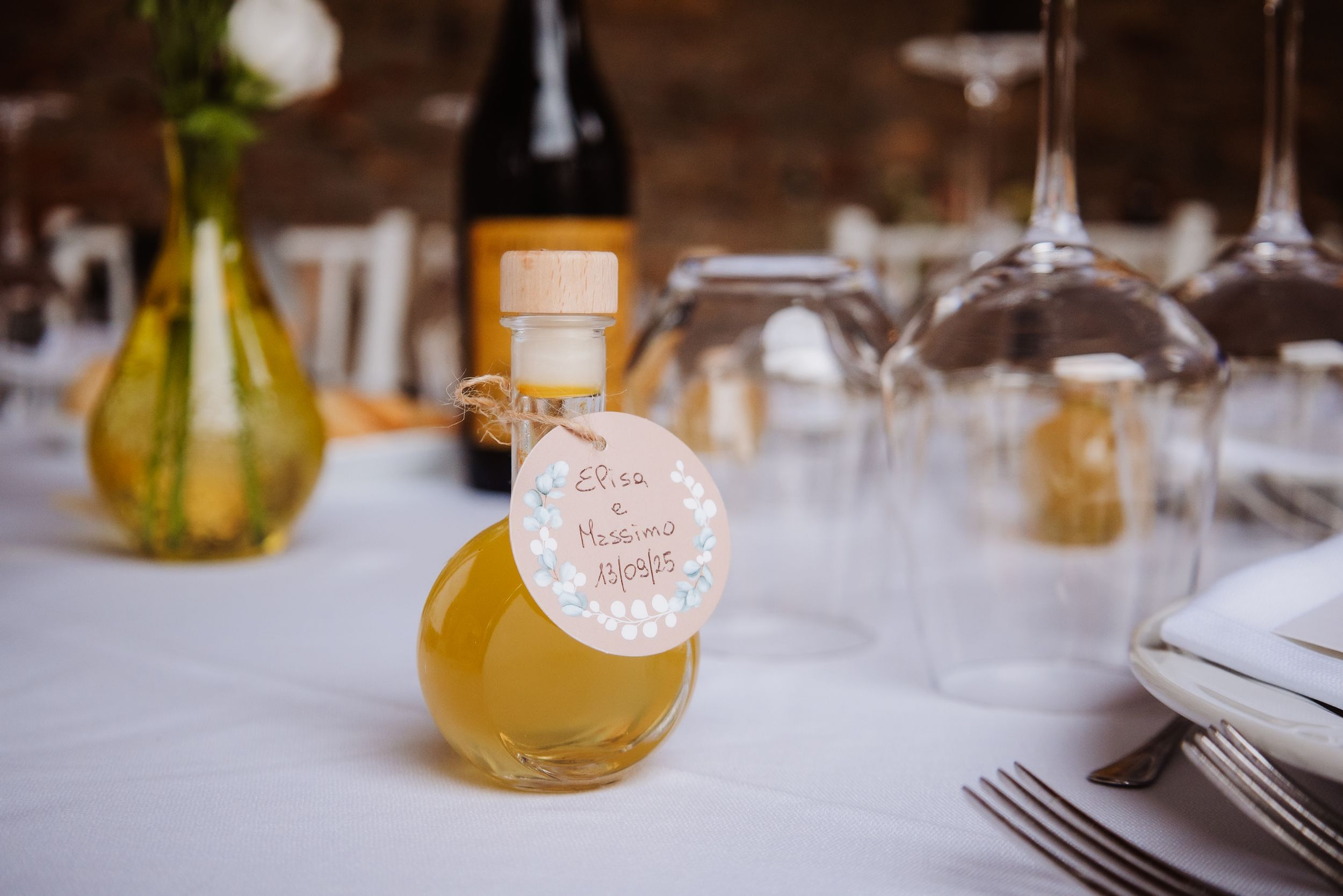 Small bottle of honey or syrup with a decorative tag sits on white tablecloth next to wine glasses and table settings.
