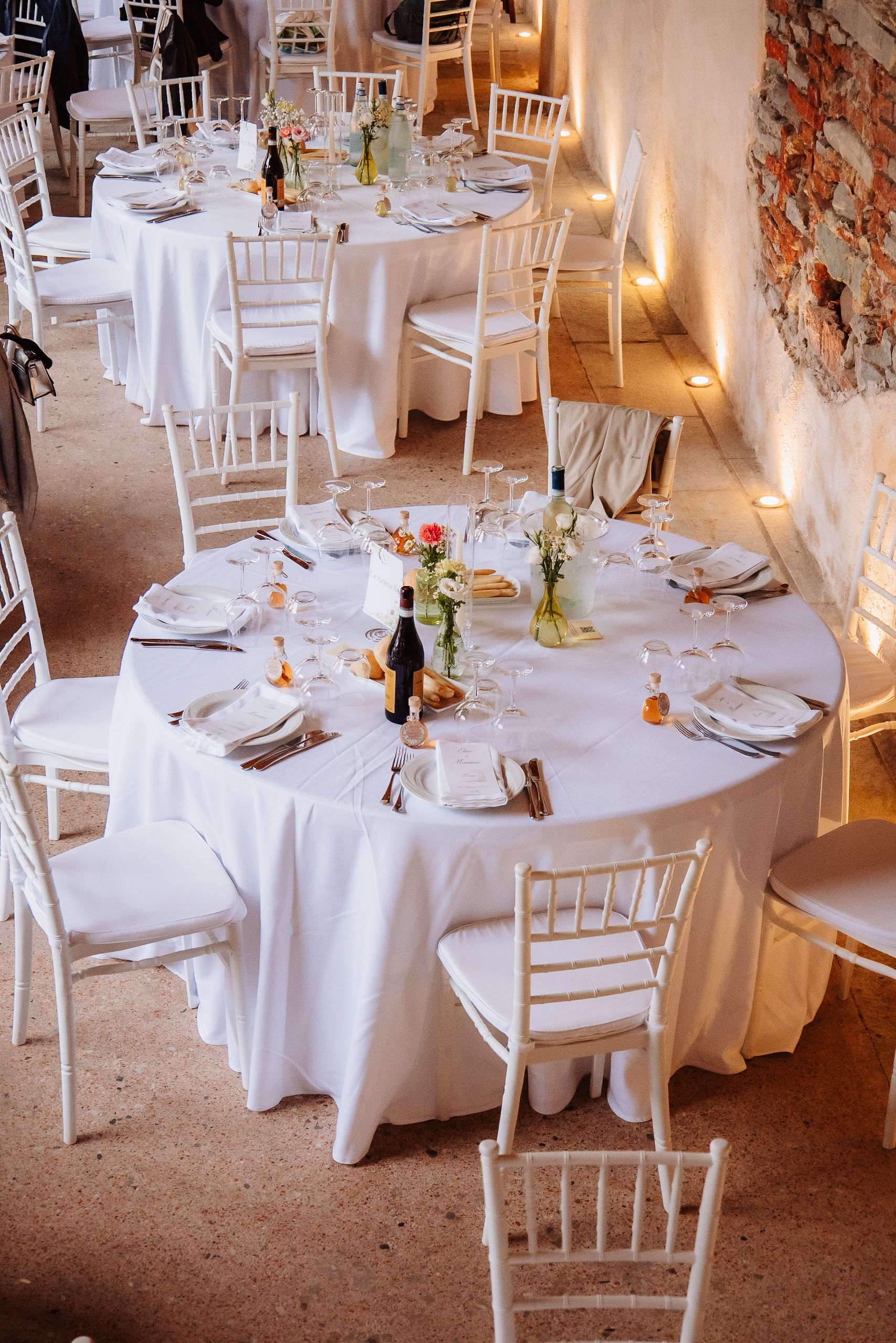Elegant wedding reception tables set with white linens, chiavari chairs, and wine bottles in a rustic brick venue.