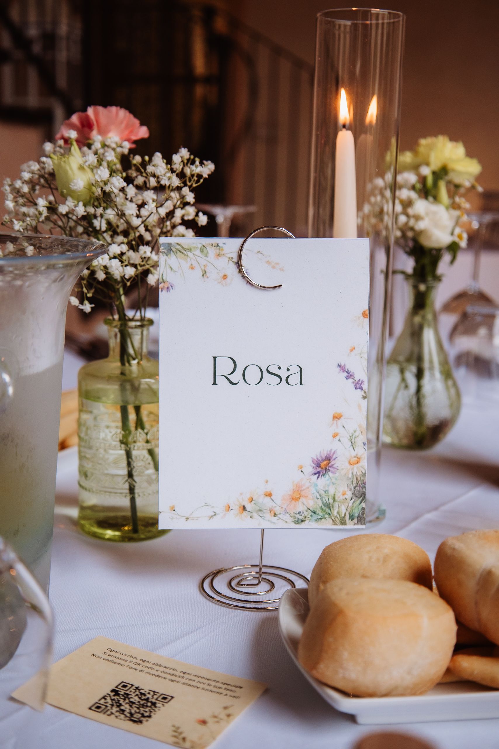 Wedding reception table setting with name card 'Rosa', delicate baby's breath flowers in mason jars, and fresh bread rolls.