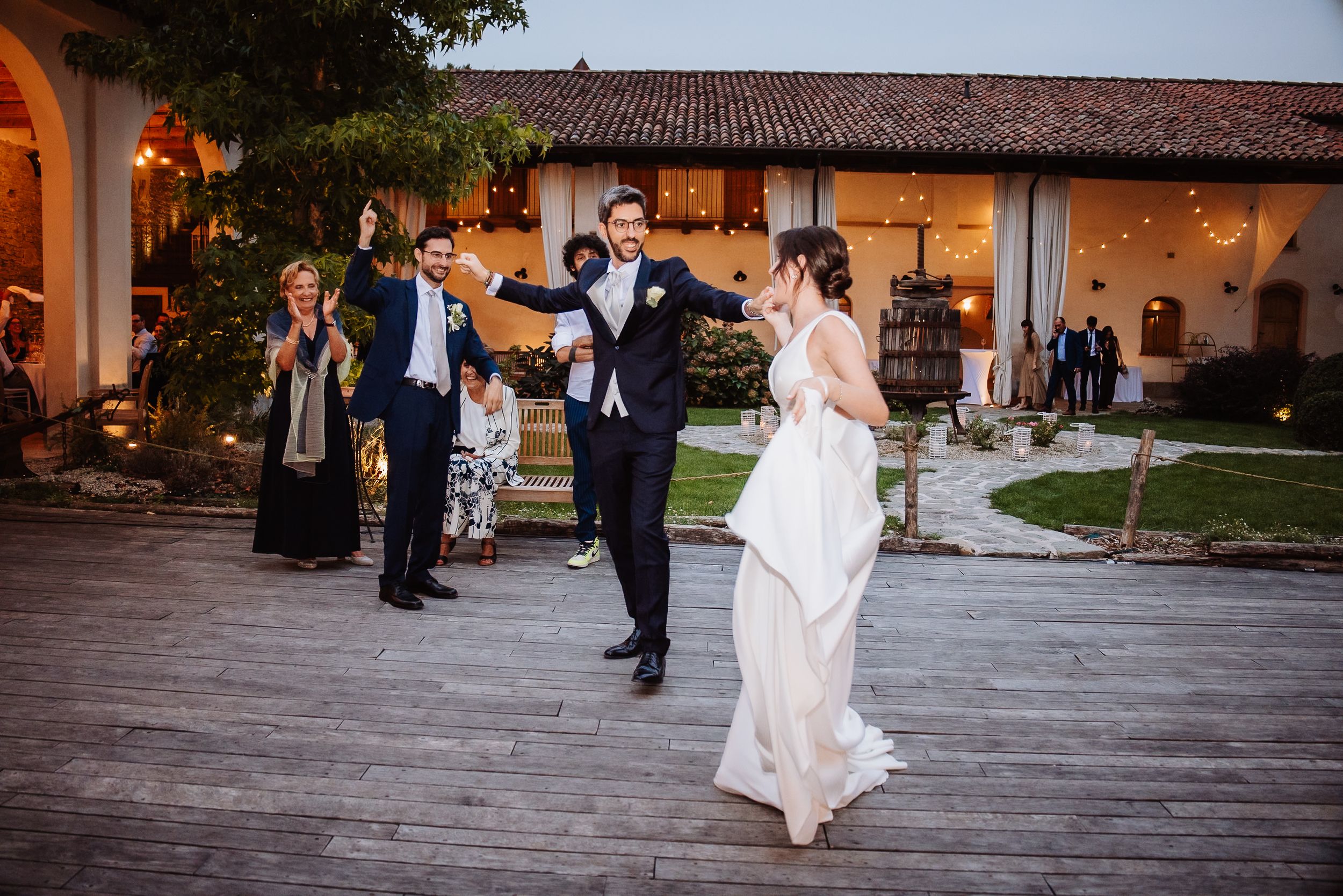 Wedding guests dance and celebrate on an outdoor stone patio at a Spanish-style venue during sunset festivities.
