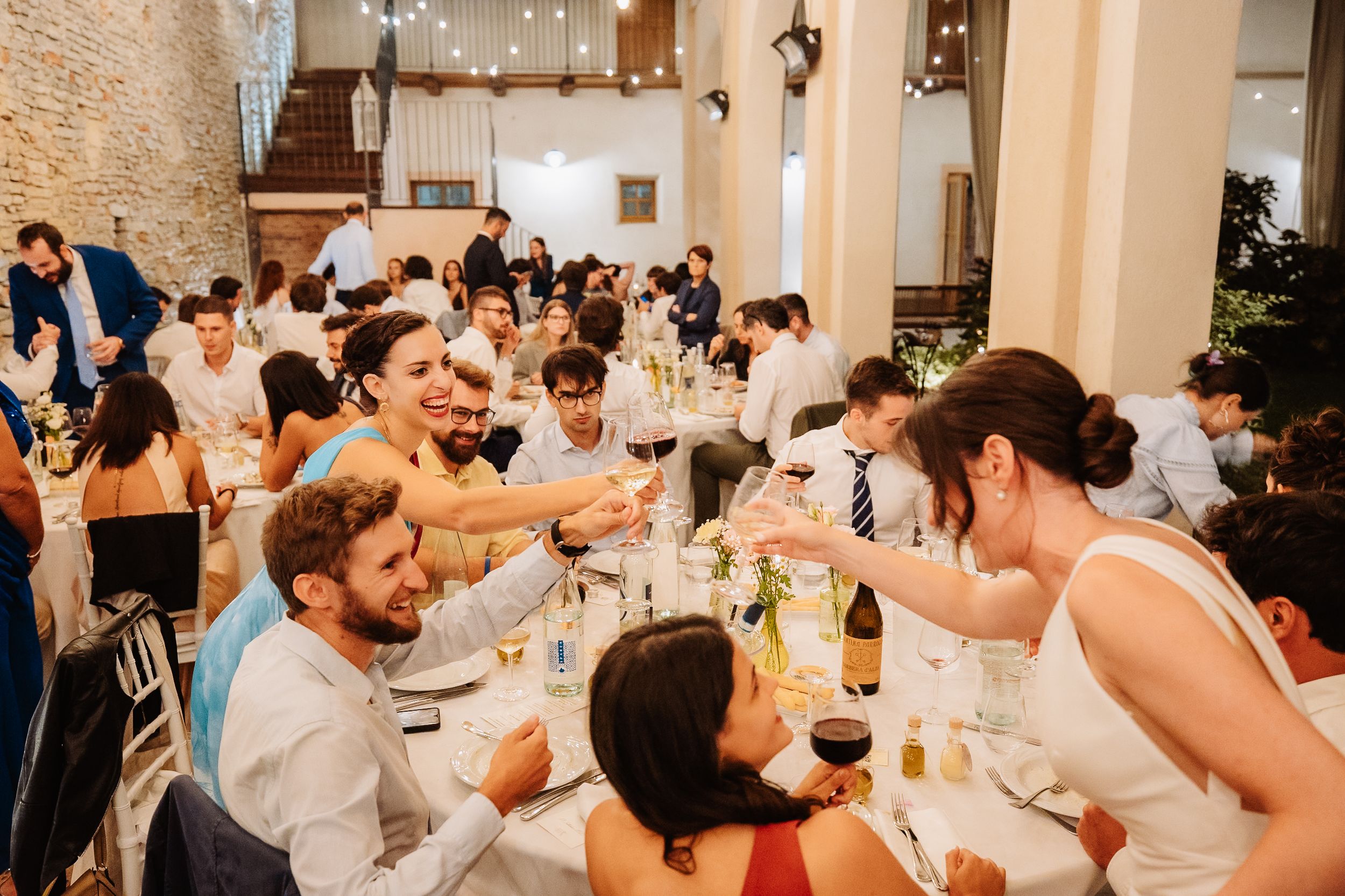 Wedding reception guests cheering and toasting at long tables decorated with string lights and stone walls.