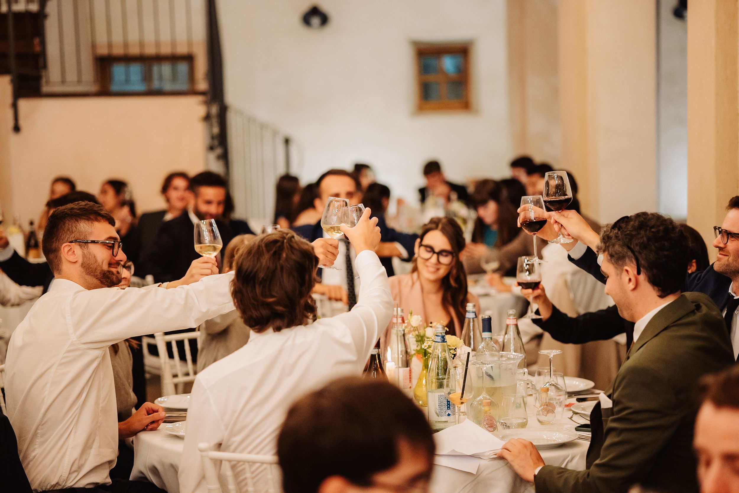 Wedding guests raise their glasses in celebration during a reception dinner in an elegant indoor venue.