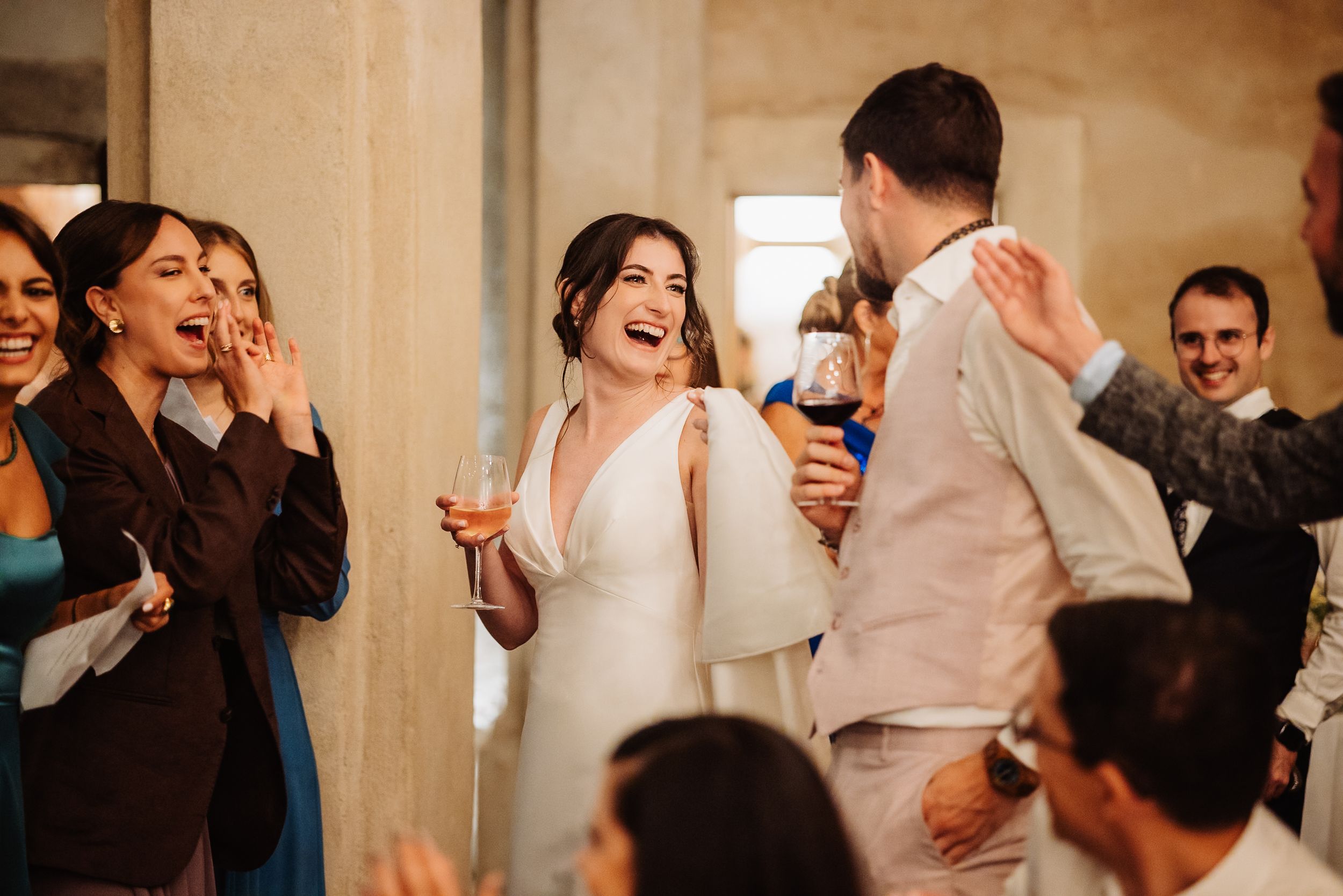 Wedding guests laugh and celebrate with drinks in hand during a joyful reception moment.