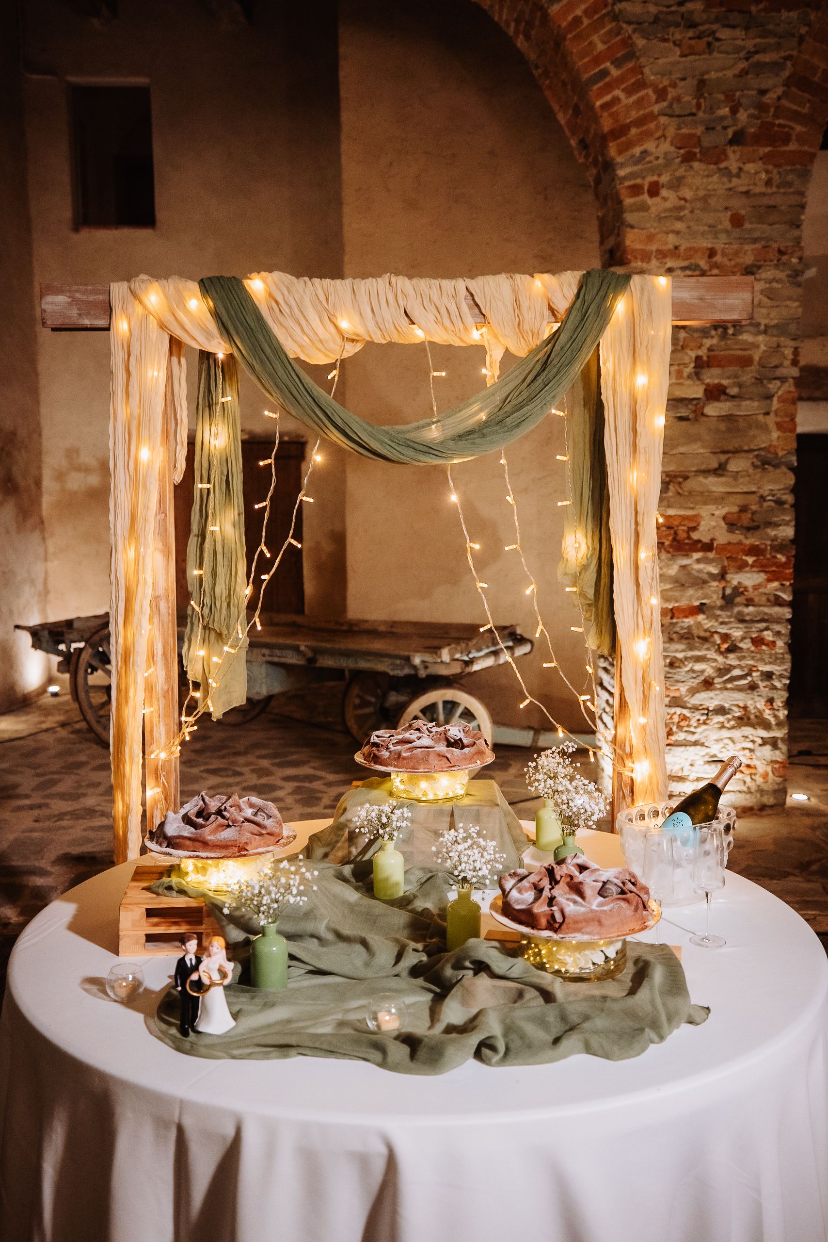 Rustic wedding dessert table with string lights, draped fabric, and cakes displayed under an illuminated wooden arch.