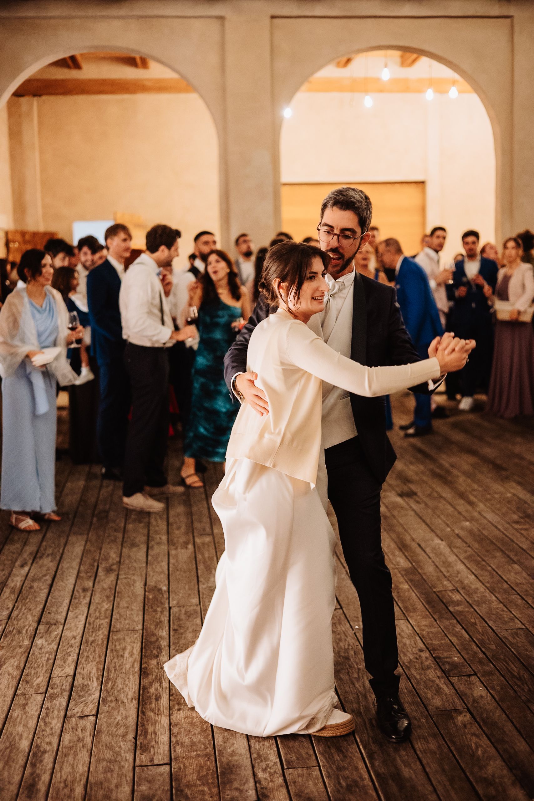 A couple shares a romantic dance together on a rustic wooden floor at their wedding reception with guests in background.