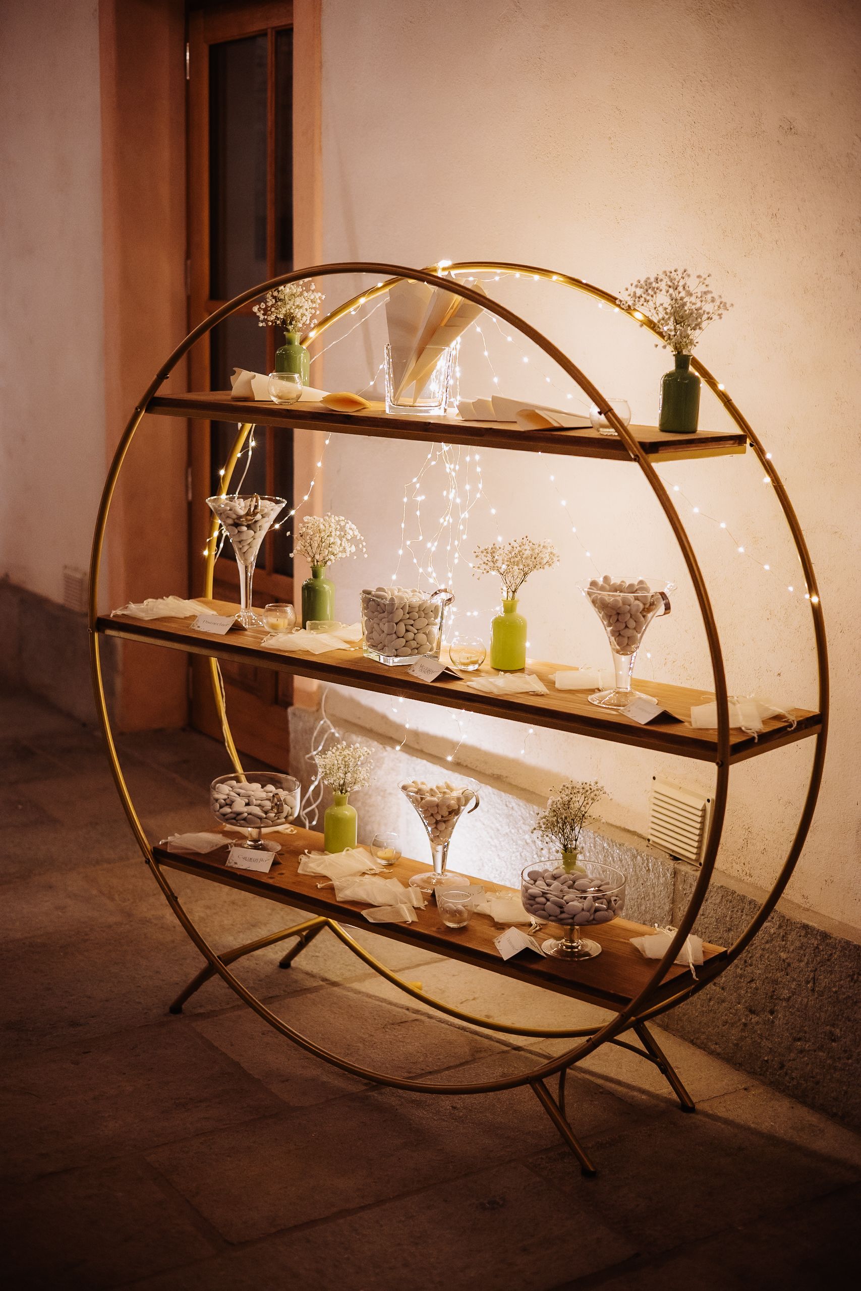 Elegant circular gold metal shelf display with wooden tiers showcasing glassware and small flower vases in warm lighting.