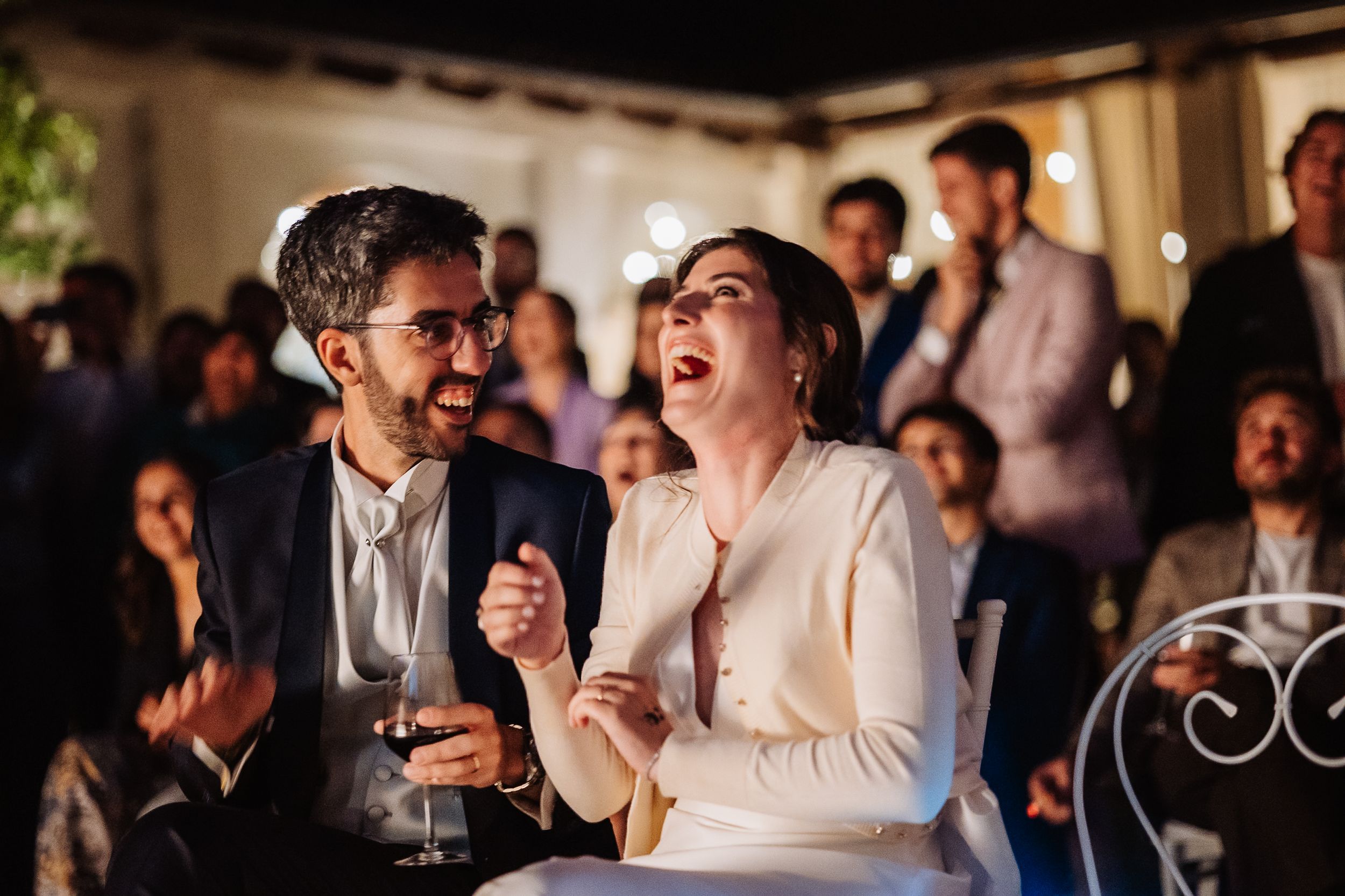 Wedding guests laugh and celebrate during a joyous reception moment under warm lighting.