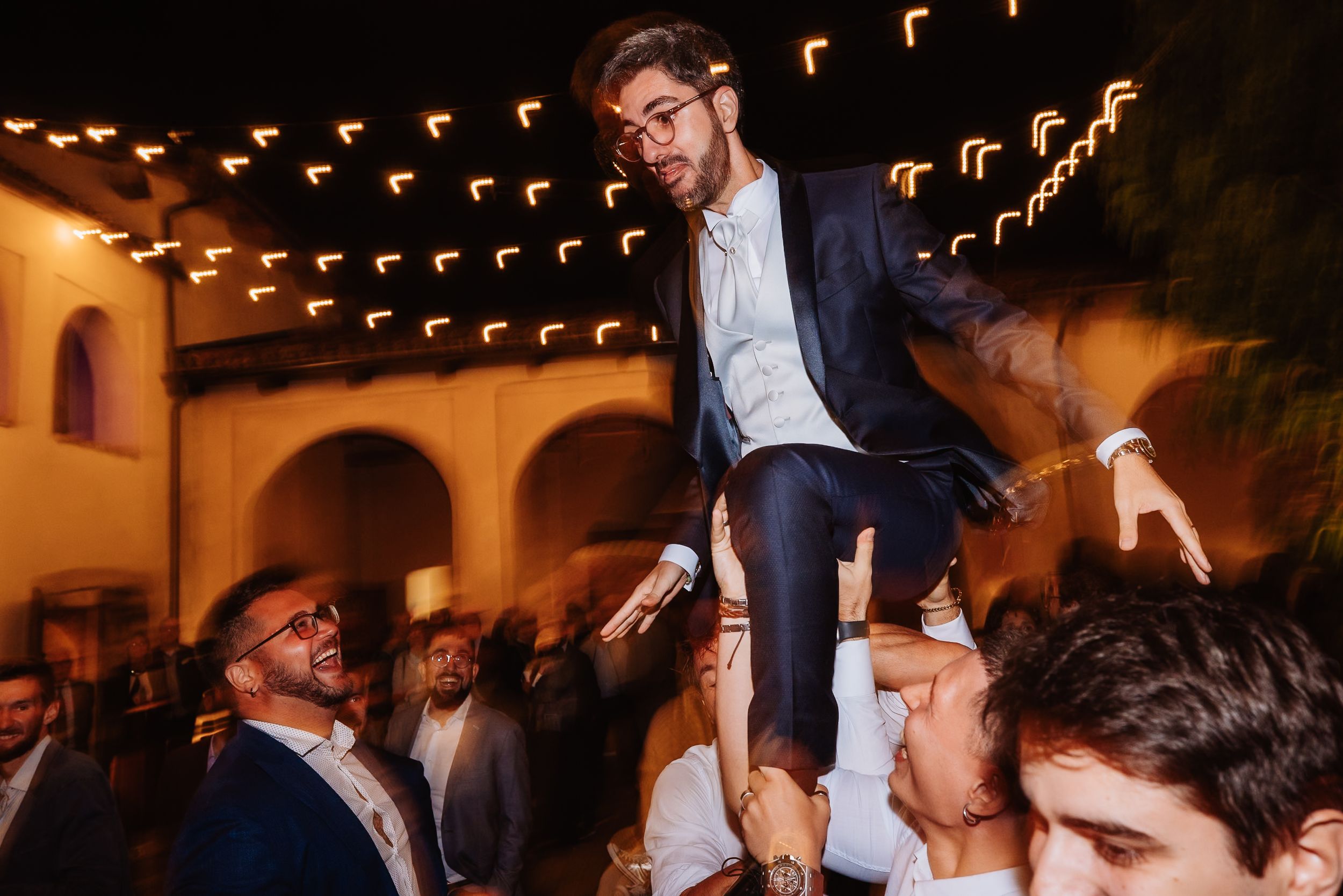 Wedding guests dance and celebrate on the dance floor under warm string lights at an evening reception.