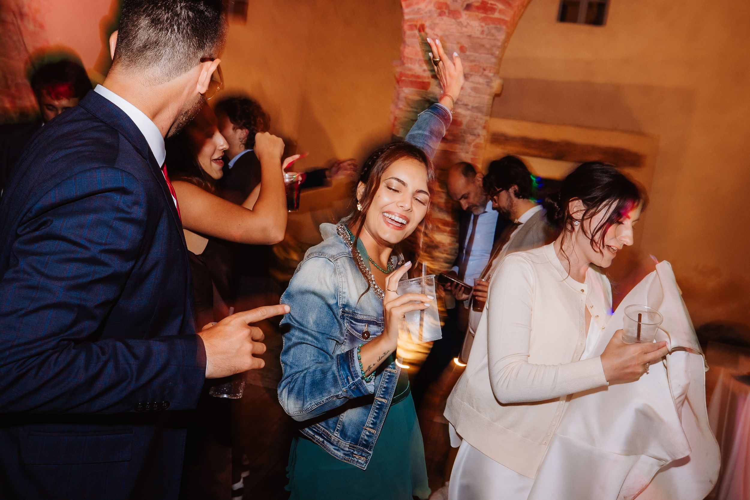 Wedding guests enjoy drinks and dance together at a lively reception with warm amber lighting.