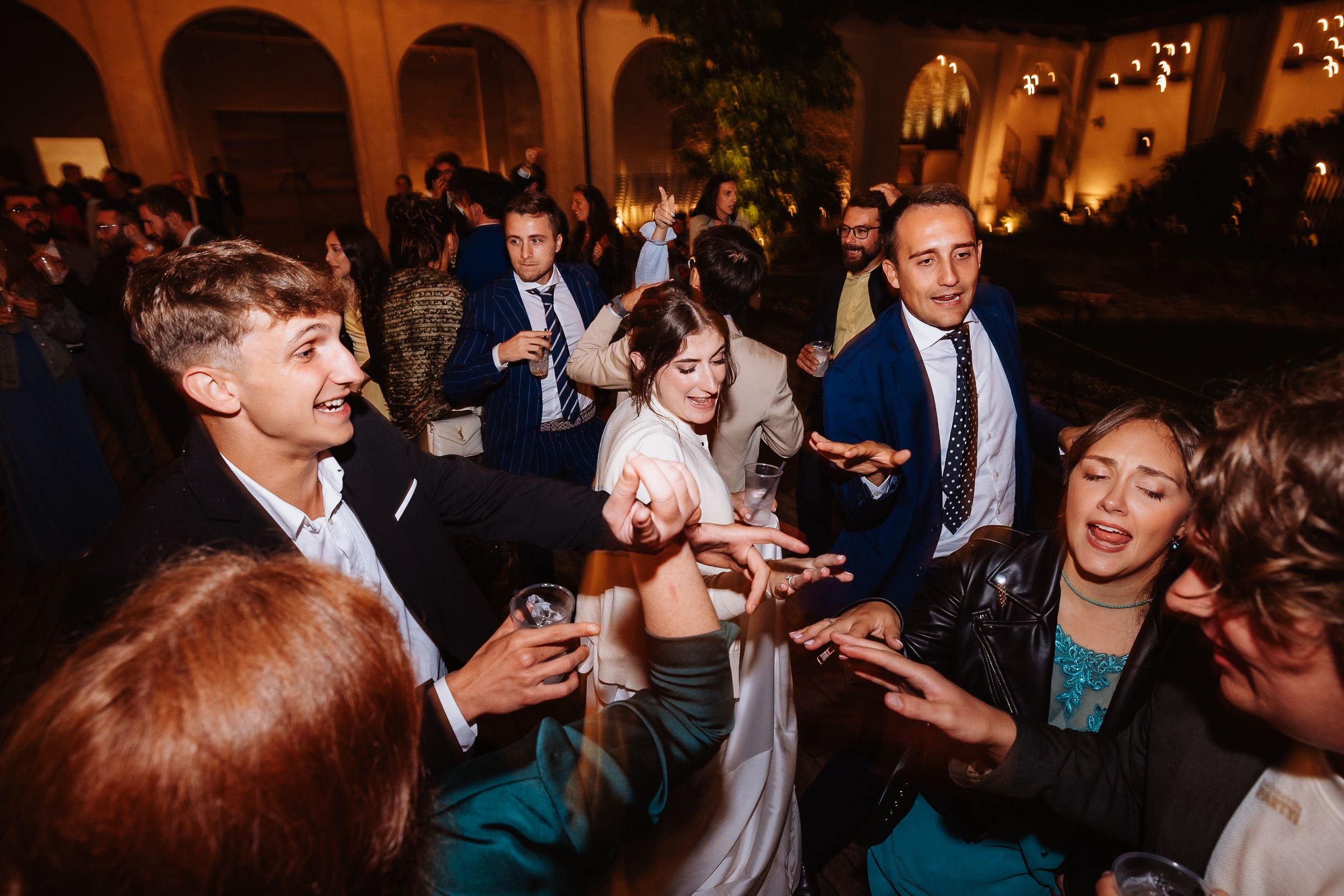 Wedding guests dance and celebrate together on a dimly lit dance floor at an evening reception party.