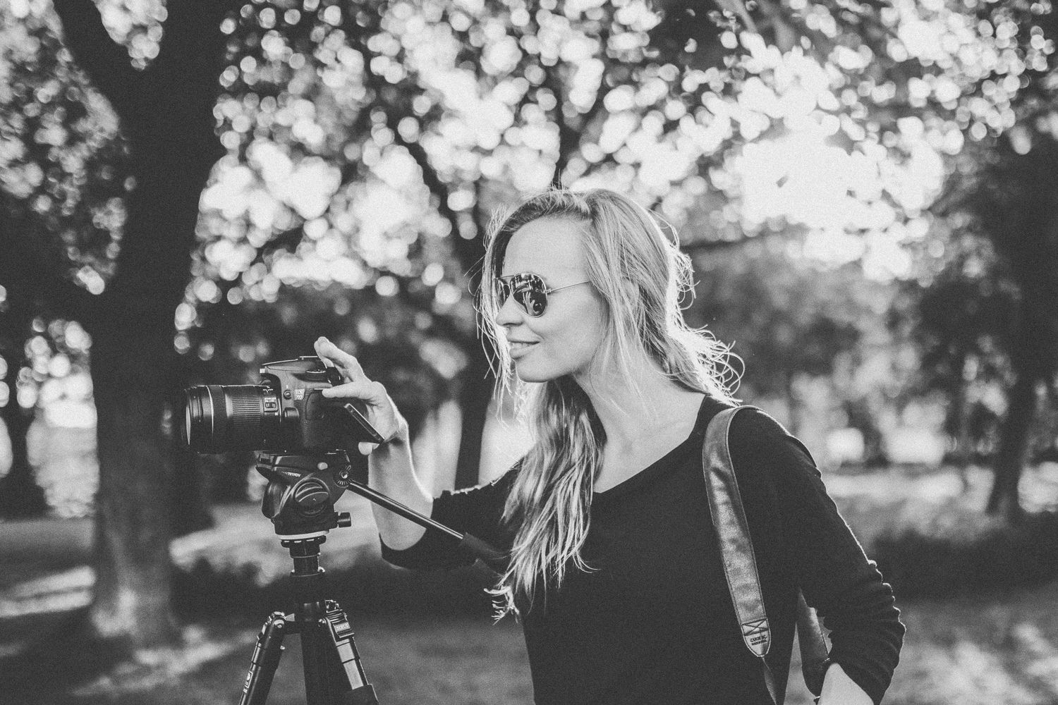 A photographer in black clothing uses a tripod mounted camera in a sunlit outdoor setting.