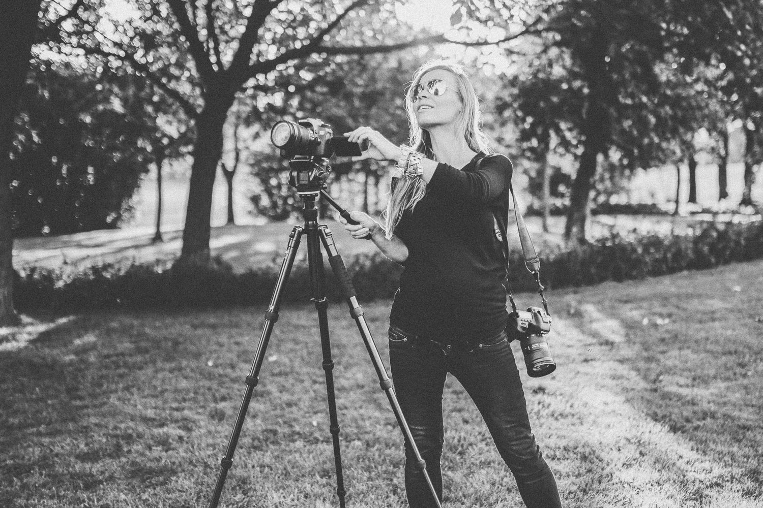 A photographer in black clothing stands next to a tripod camera outdoors in a black and white photograph.