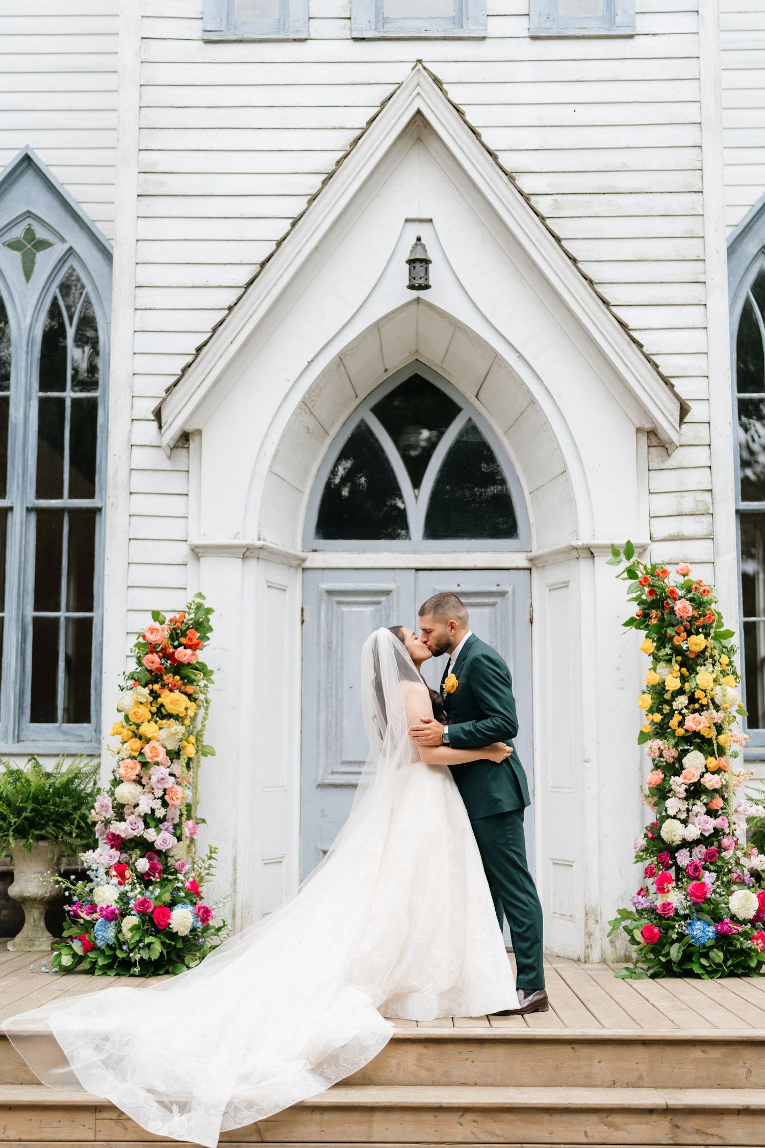 A romantic wedding scene at a white chapel entrance with gothic windows and beautiful floral arrangements on either side.