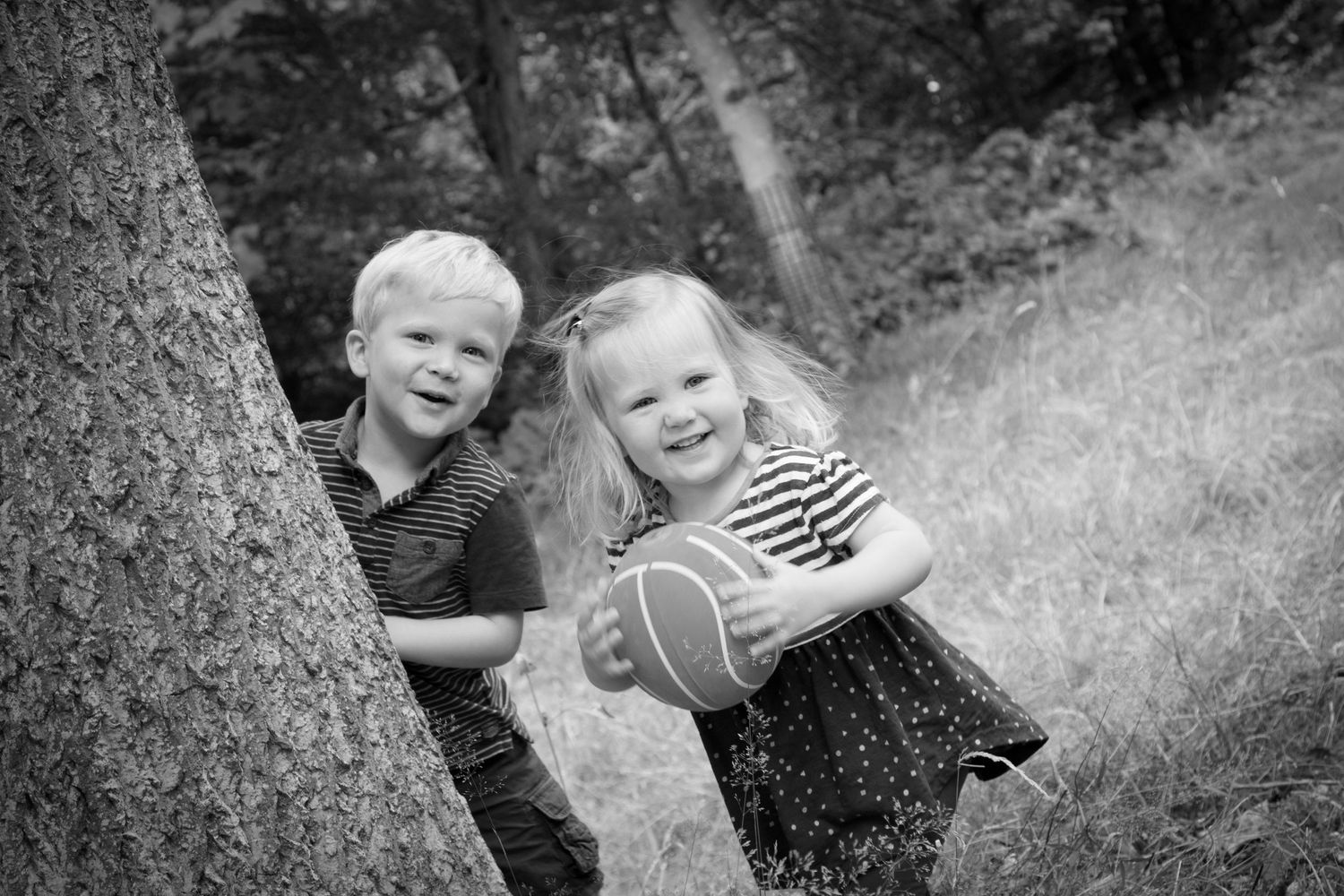 Black and white nursery photo of siblings peeking out behind a tree playing peekaboo and laughing.