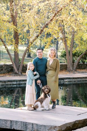A family stands with their fluffy brown and white dog on a concrete platform by a pond surrounded by autumn trees.
