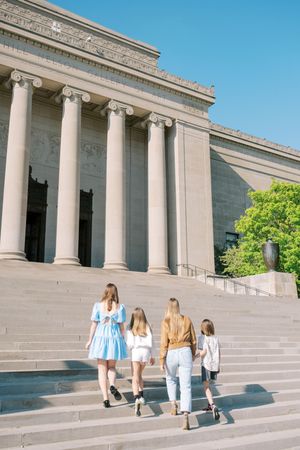 Group of visitors walking up the grand stone steps of a neoclassical museum building with white columns on a sunny day.