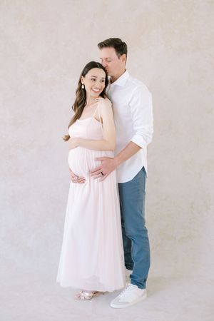 A couple in casual attire share tender moments against a light backdrop in a series of romantic maternity photos.