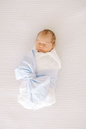 A sleeping newborn baby swaddled in white and light blue blankets on a textured white background.